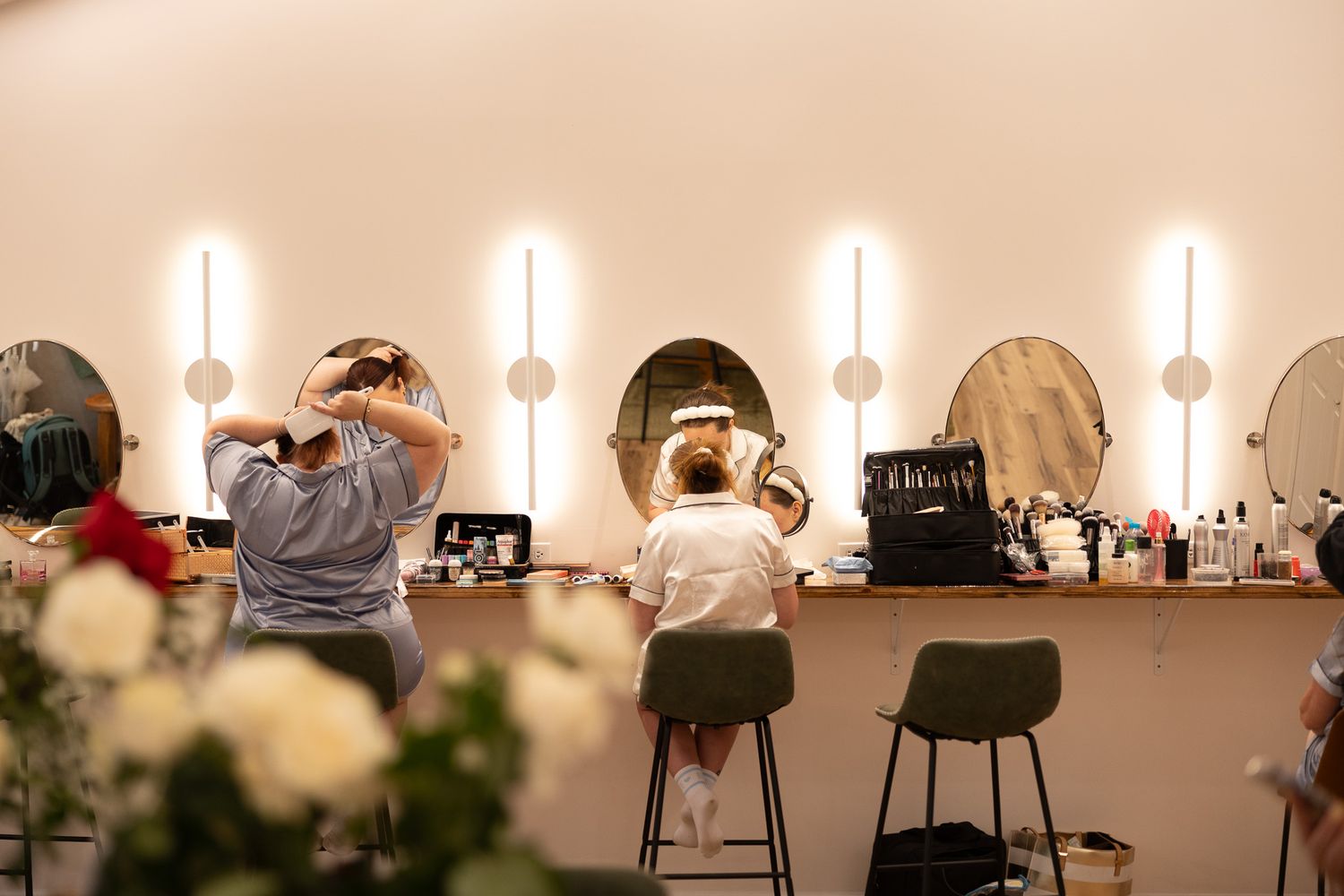 Two women sit at a vanity mirror station with bright lights in a modern beauty salon getting hair and makeup done.