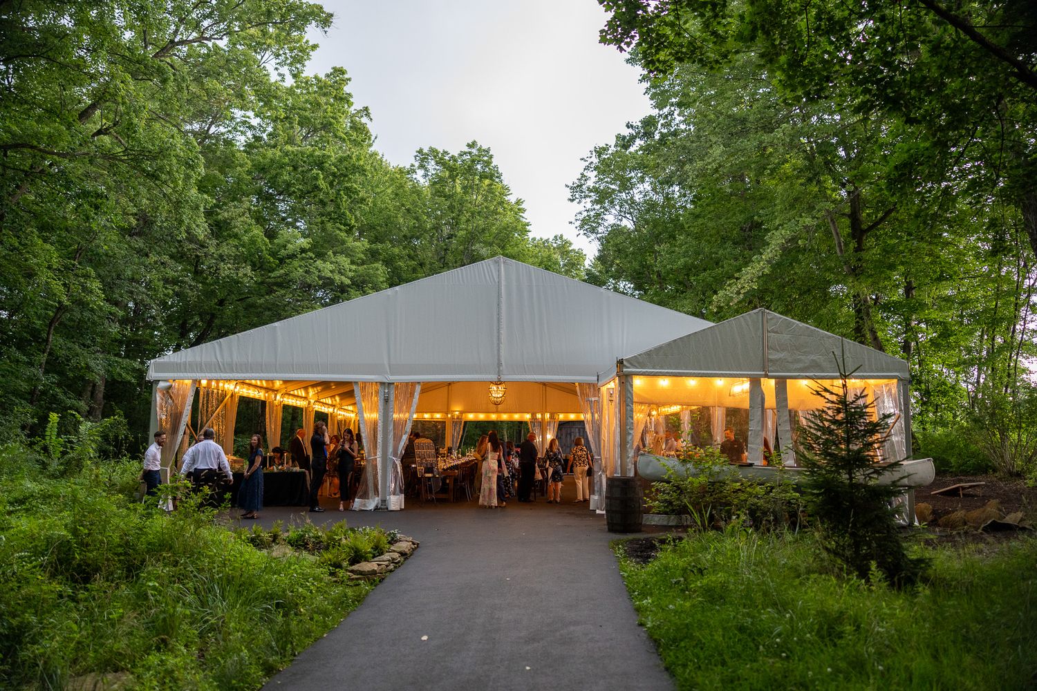 Elegant outdoor wedding tent glowing with warm lights surrounded by lush green trees at dusk.