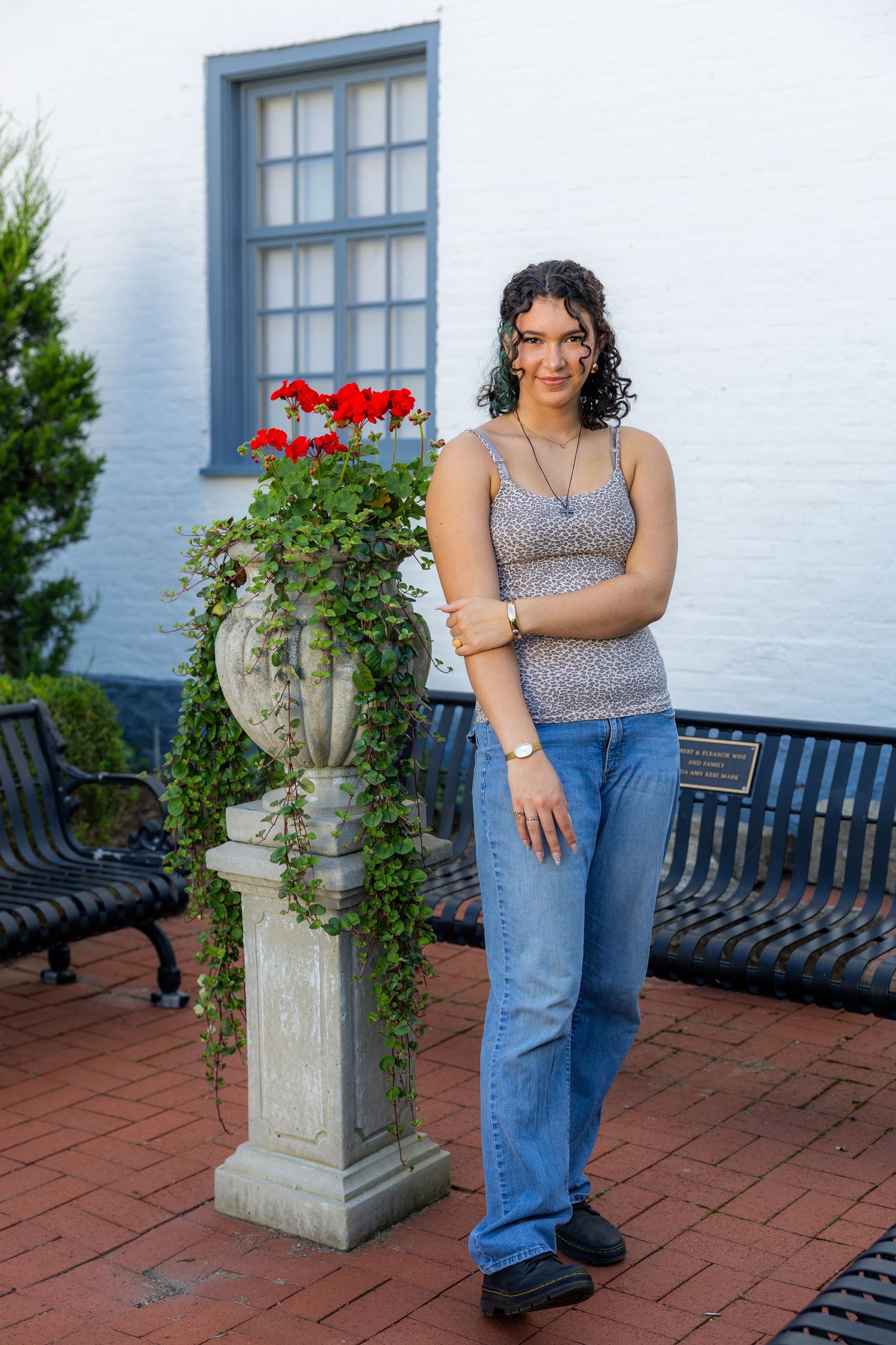 Young woman in jeans and tank top posing beside a stone urn planter with red geraniums outside a white building.