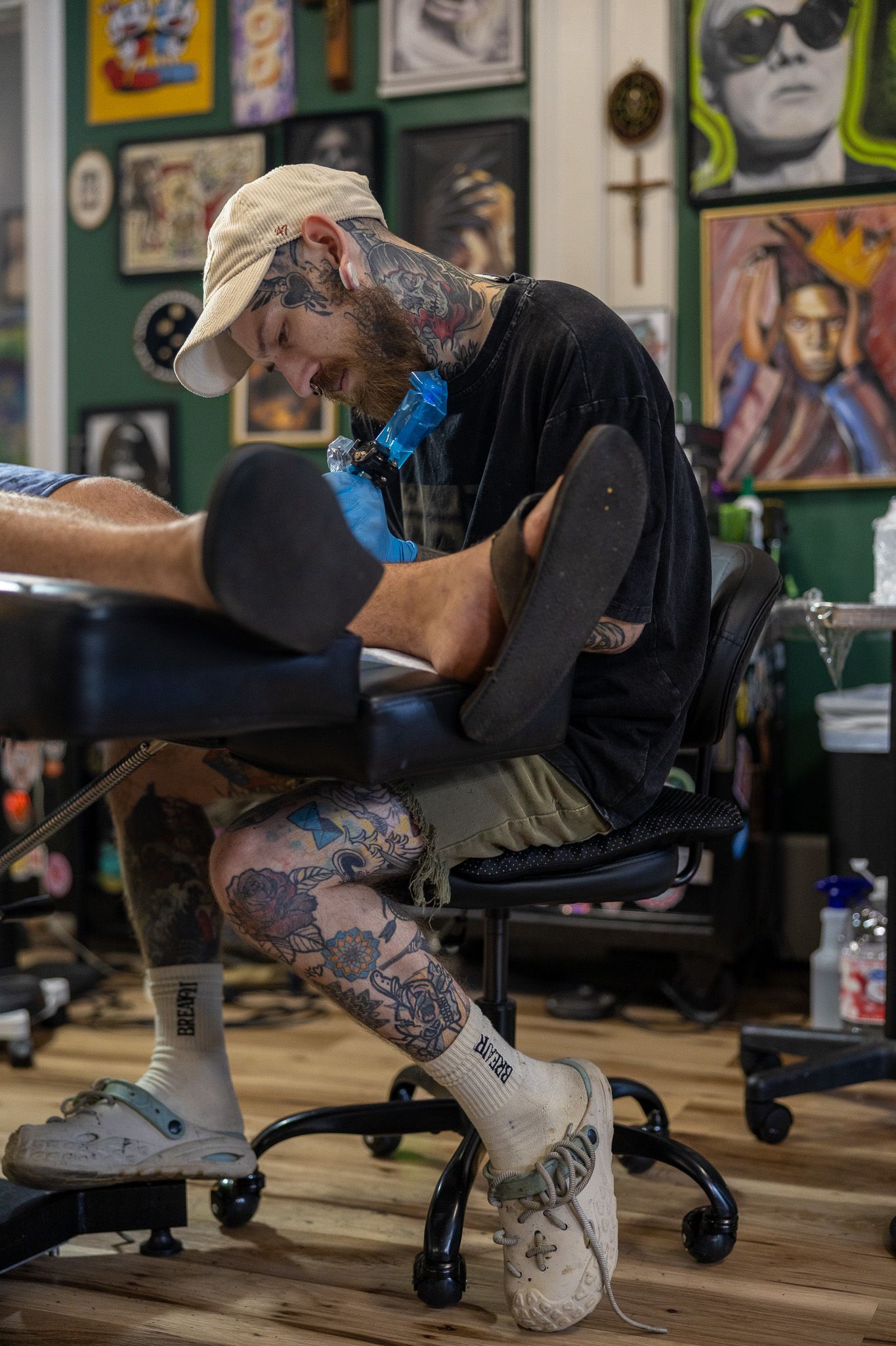 Tattooed artist tattooing a client's leg in a vibrant tattoo studio filled with colorful artwork on the walls.
