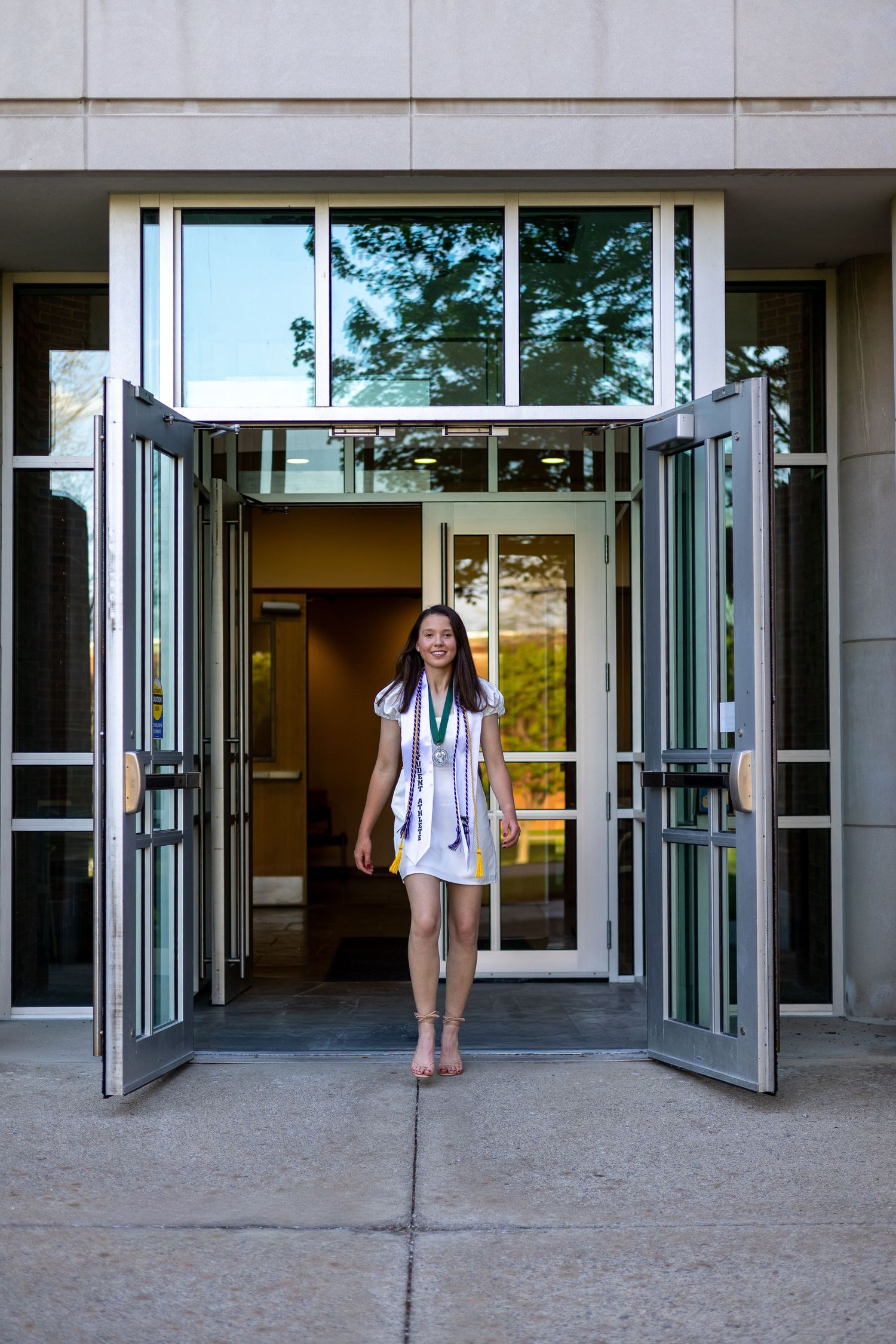 Smiling graduate in white dress walks through open doors of a modern university building.