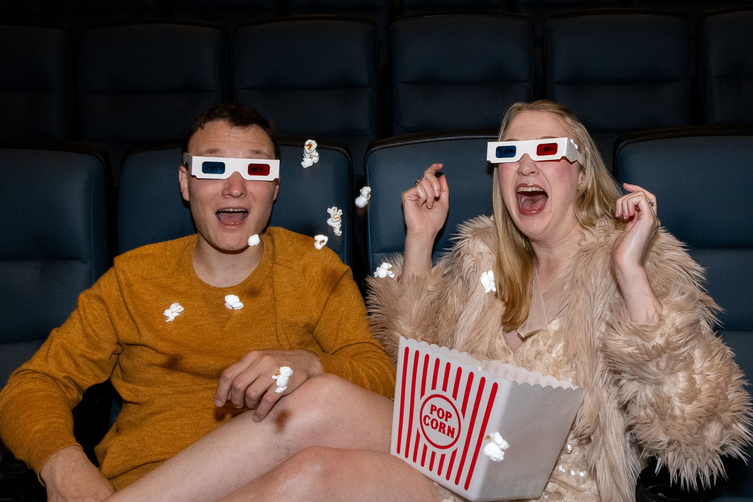 Excited couple in 3D glasses with popcorn flying at a movie theater.