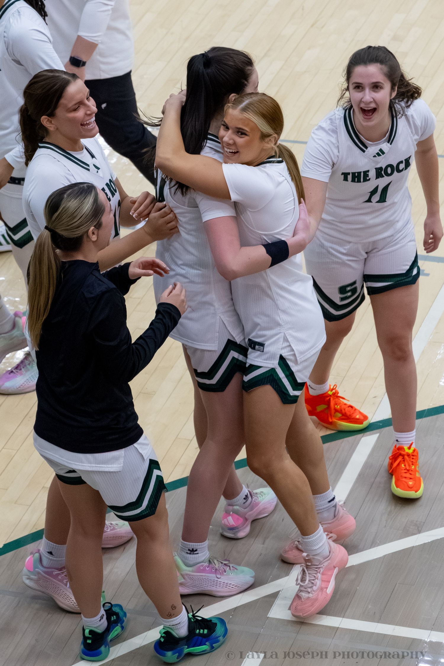 Women's basketball team in white The Rock uniforms celebrating with hugs on the court.