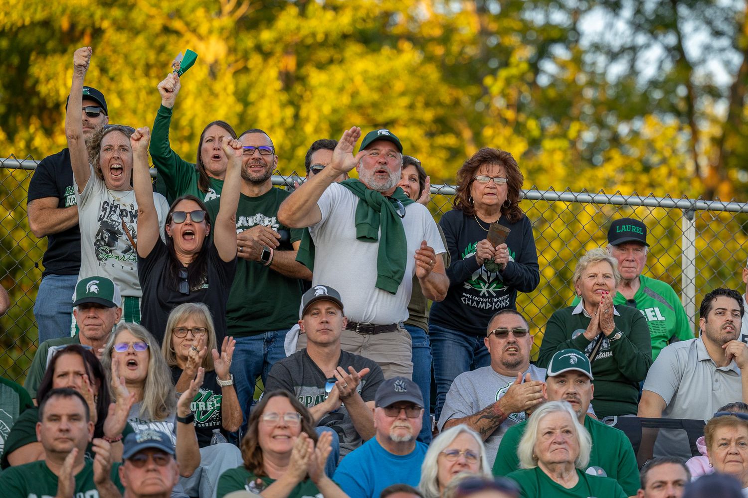 Excited fans in green and white cheer enthusiastically at an outdoor Michigan State Spartans sporting event.