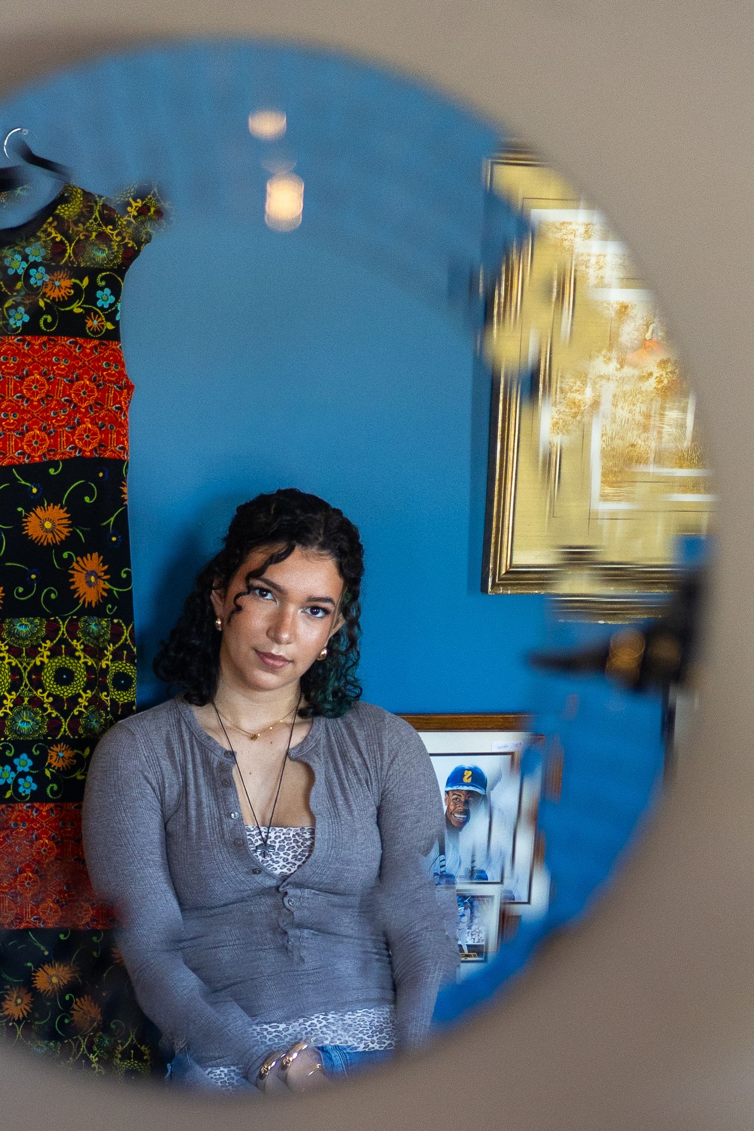 Young woman with curly hair in gray top reflected in round mirror, blue wall and colorful dress visible behind her.