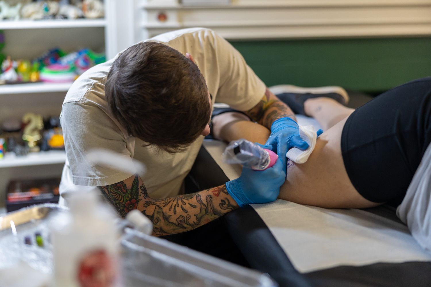 Tattoo artist leaning over and carefully tattooing a design onto a clients leg in a bright studio.