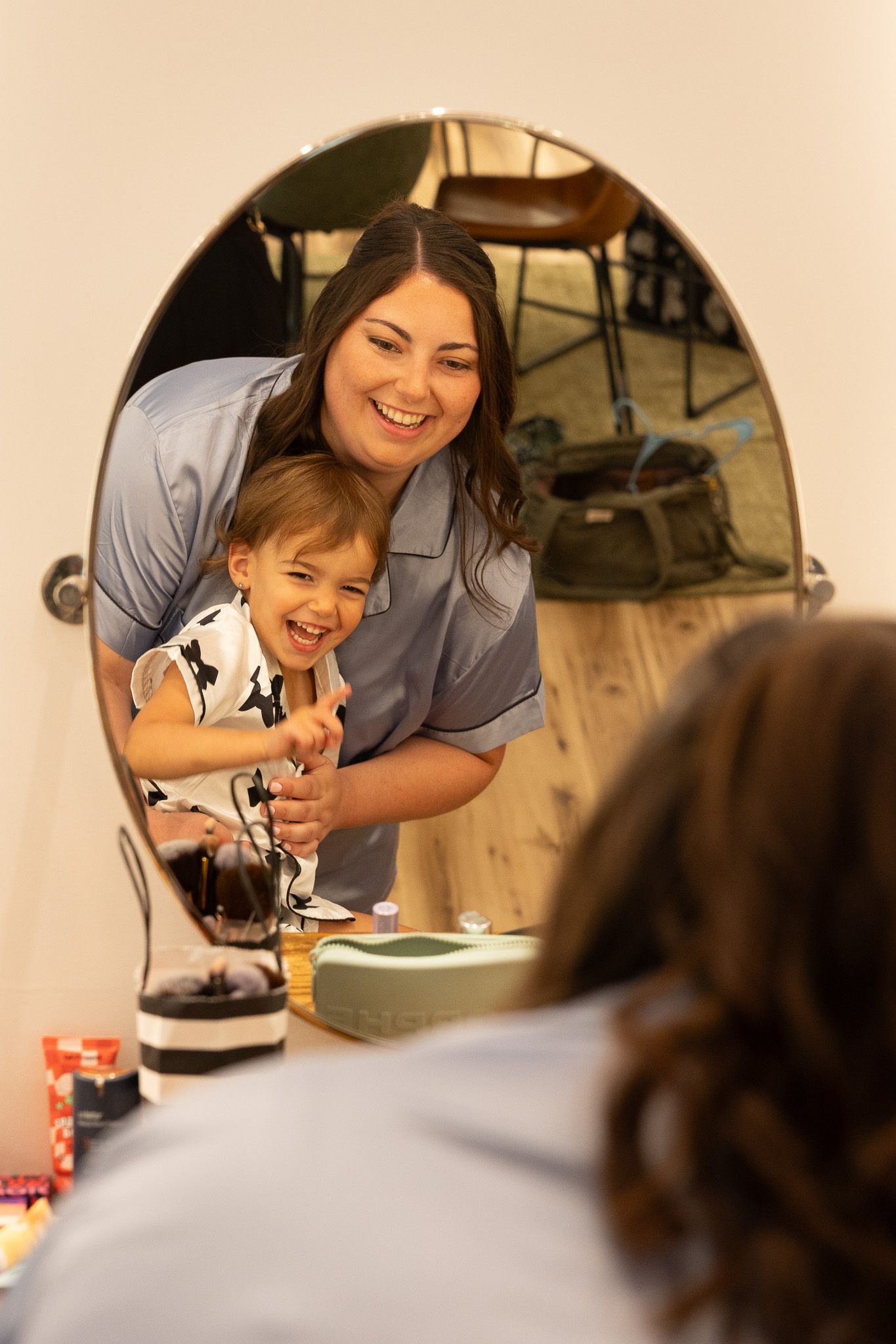 A smiling mother and young child laugh together while looking at their reflection in a round bedroom mirror.