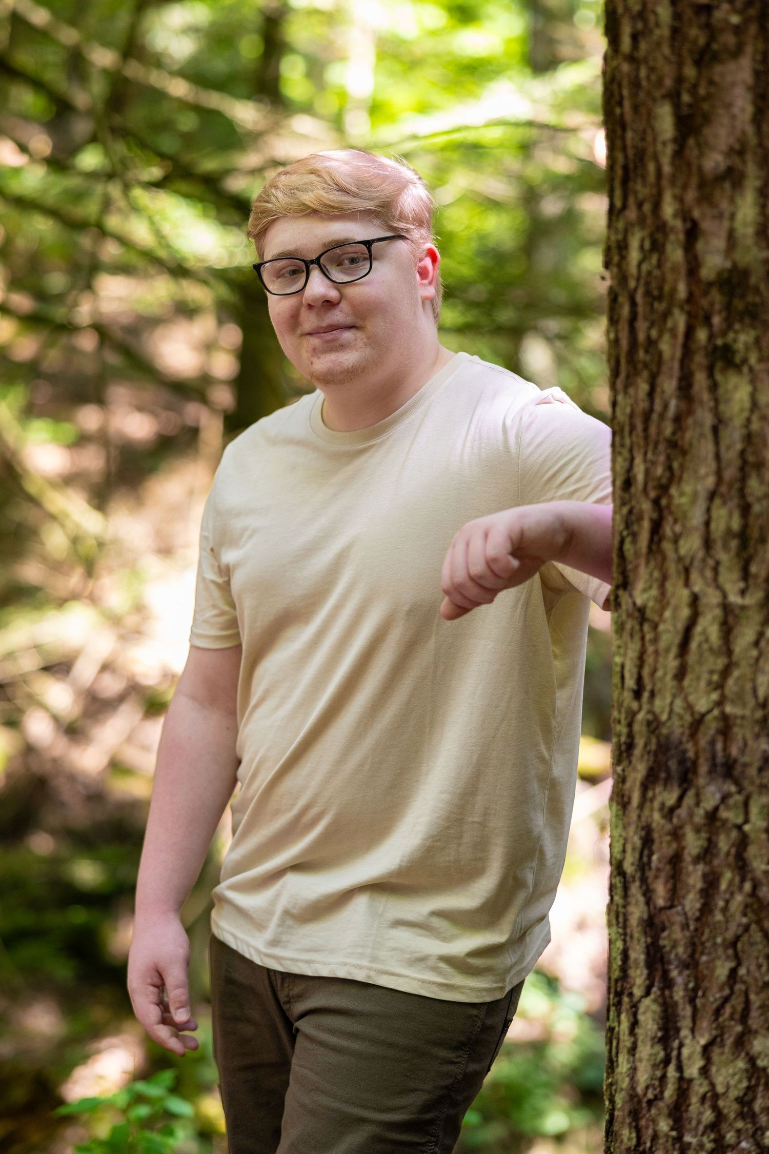 Young man with glasses in a beige t-shirt leaning against a tree in a sunlit forest.