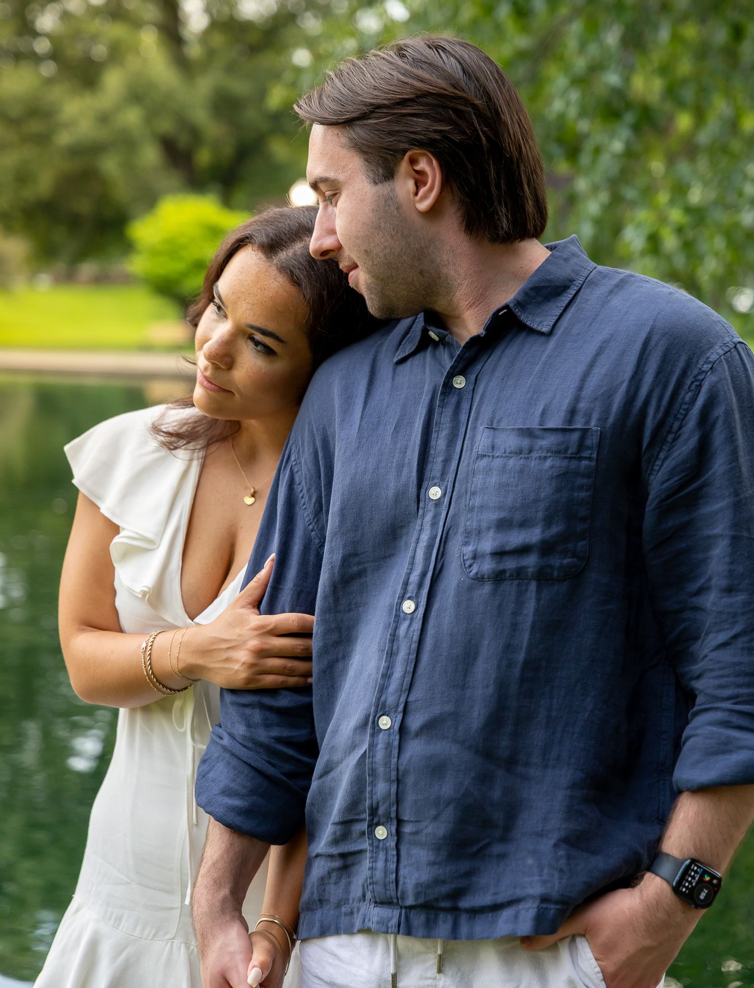 Engaged couple embracing by a serene pond in a lush green park setting.