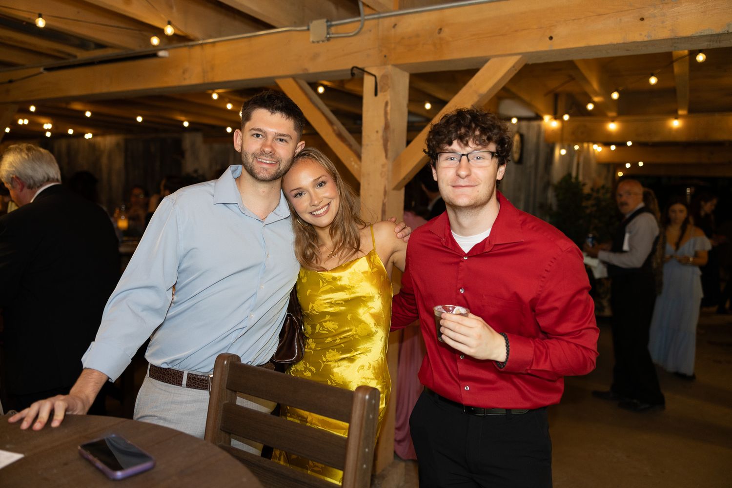 Three wedding guests smile together in a rustic barn venue with warm string lights overhead.