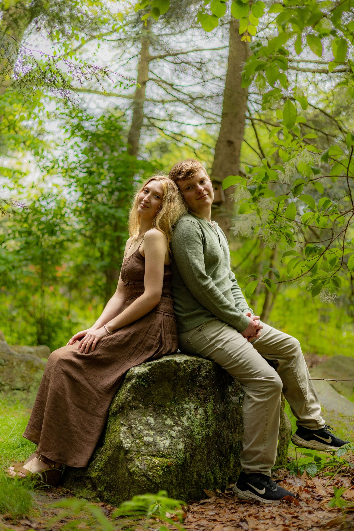 A young couple sits back-to-back on a mossy rock surrounded by lush green trees.