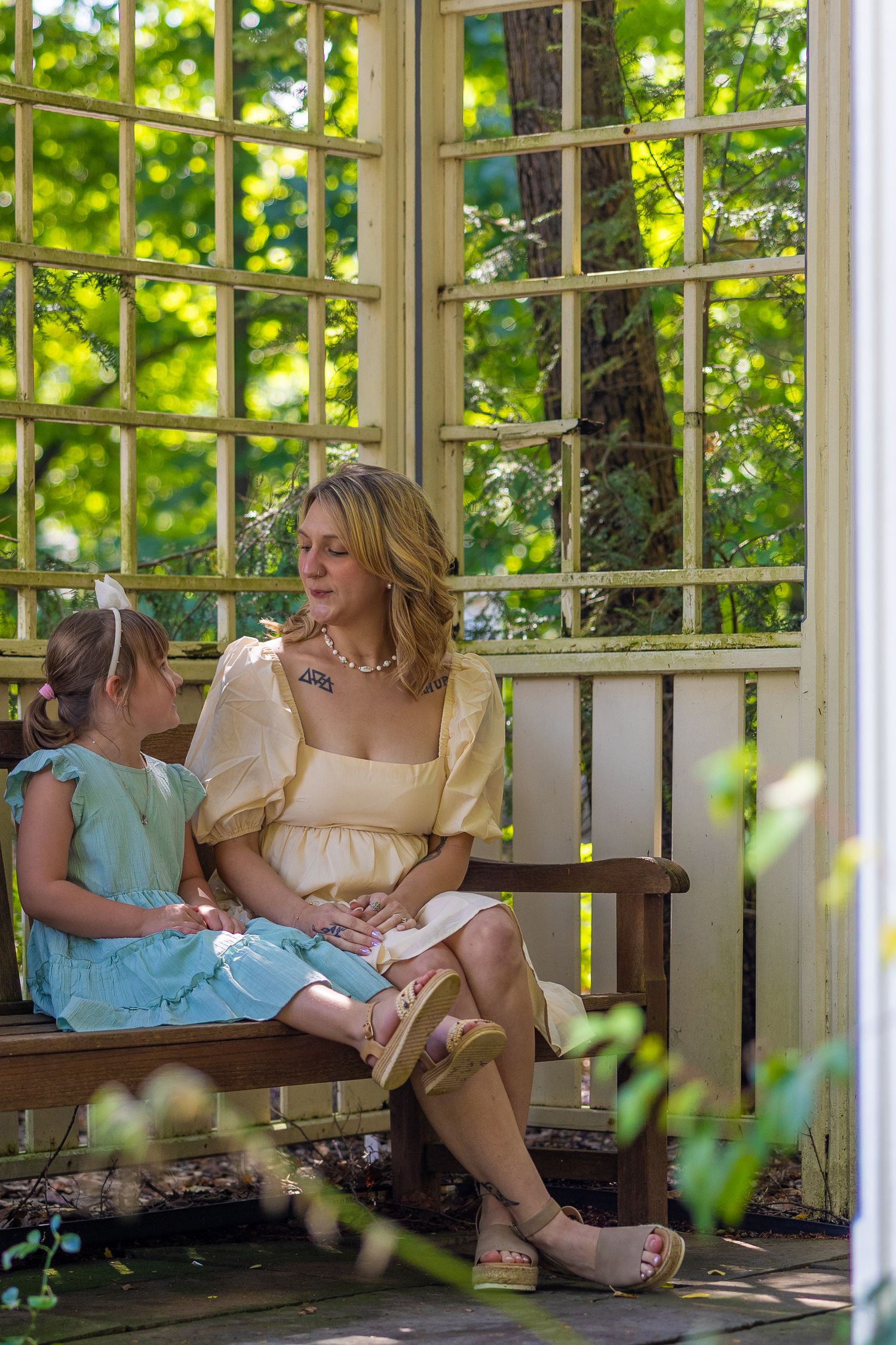 Woman in yellow dress sits with young girl in blue dress on bench in sunlit garden gazebo.