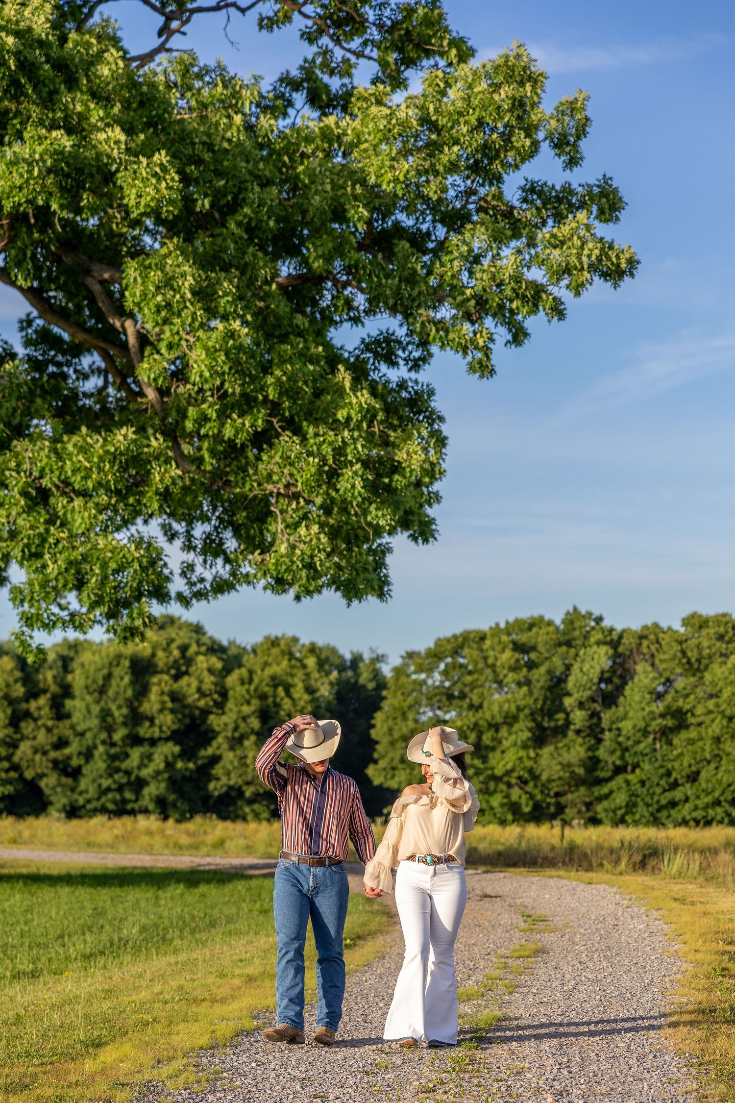 Couple in cowboy hats holding hands while walking down a gravel path surrounded by lush green trees and fields.
