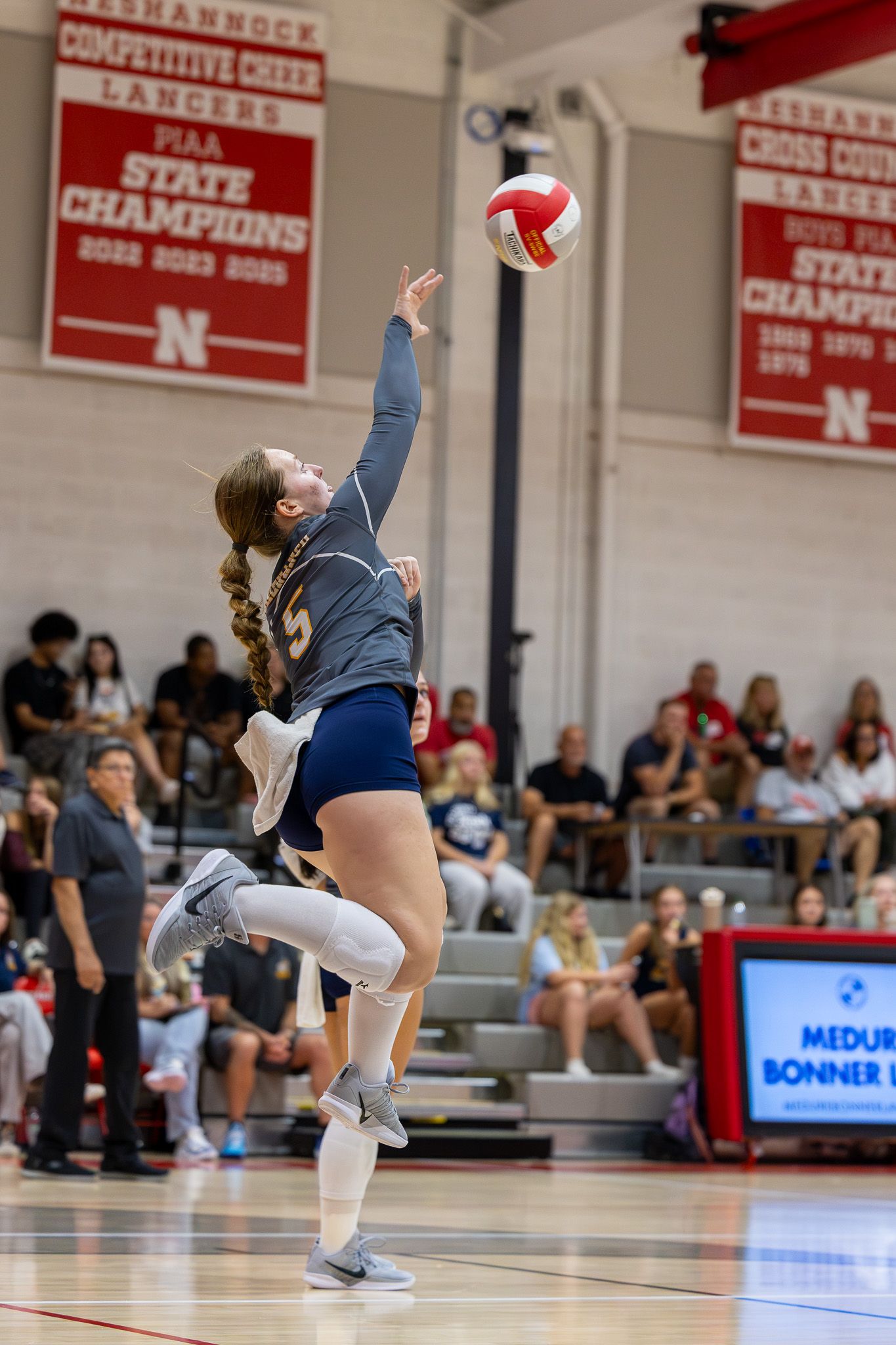 Female volleyball player leaping to serve the ball during an indoor high school match with spectators in the background.