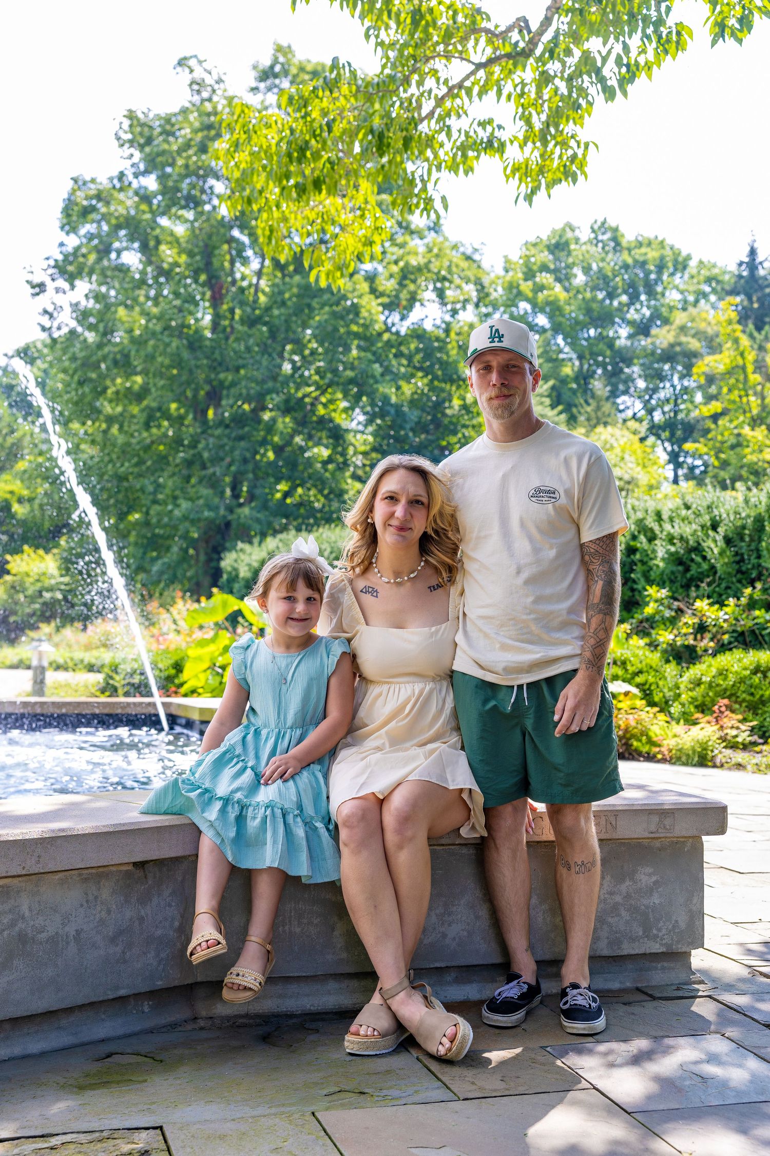 Family of three posing by a fountain in a lush green park on a sunny day, wearing summer outfits.