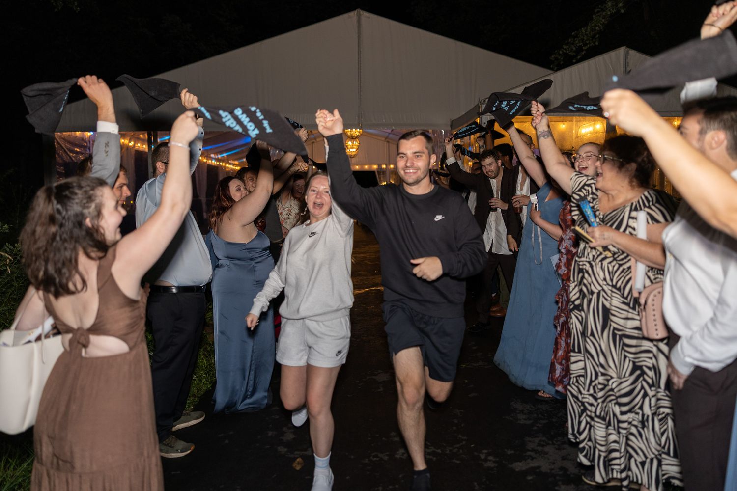 Excited crowd celebrating outdoors at night under a tent with raised hands and umbrellas.