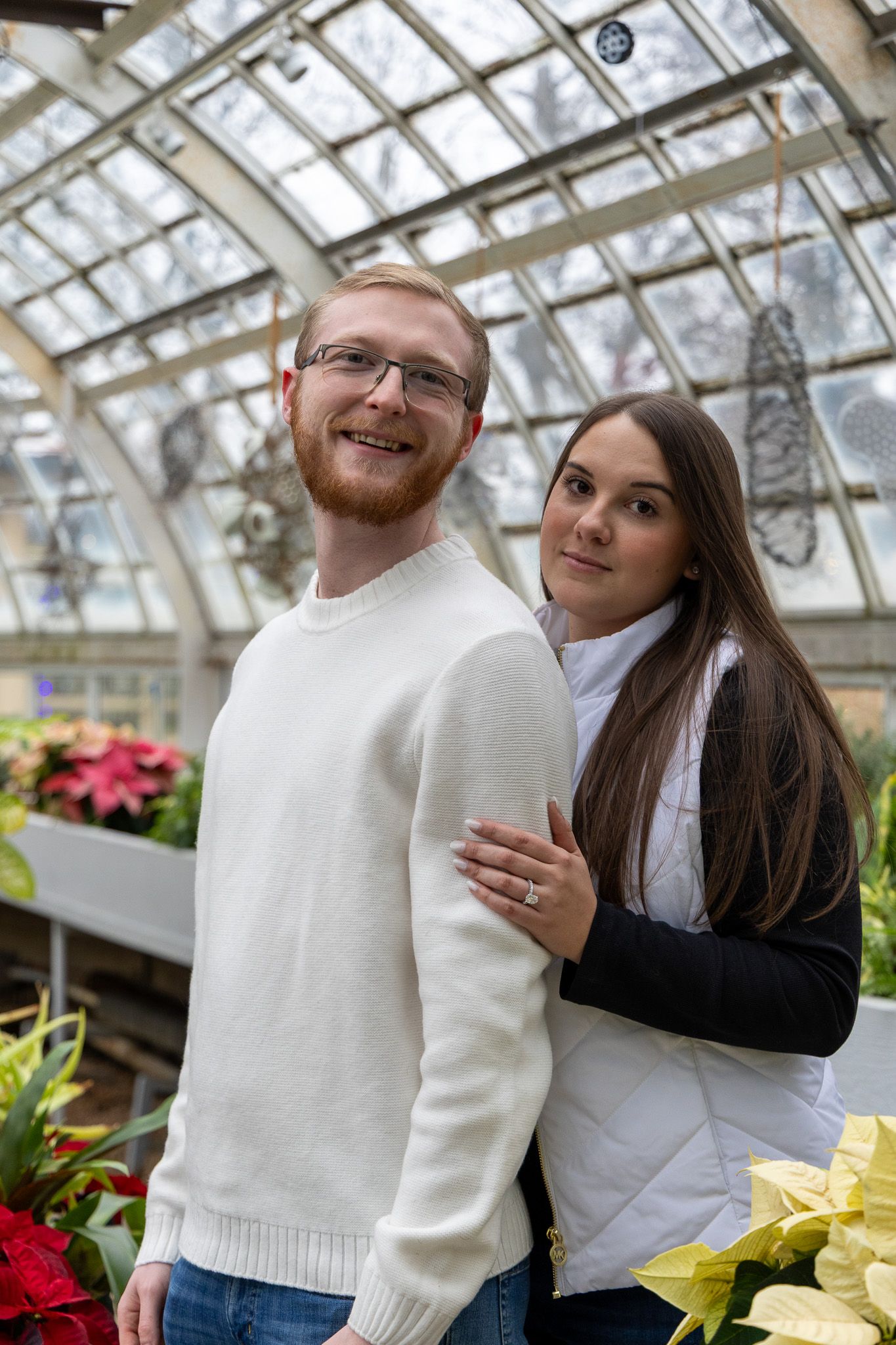 A smiling couple poses in a glass greenhouse surrounded by colorful poinsettias, man in white sweater with glasses.