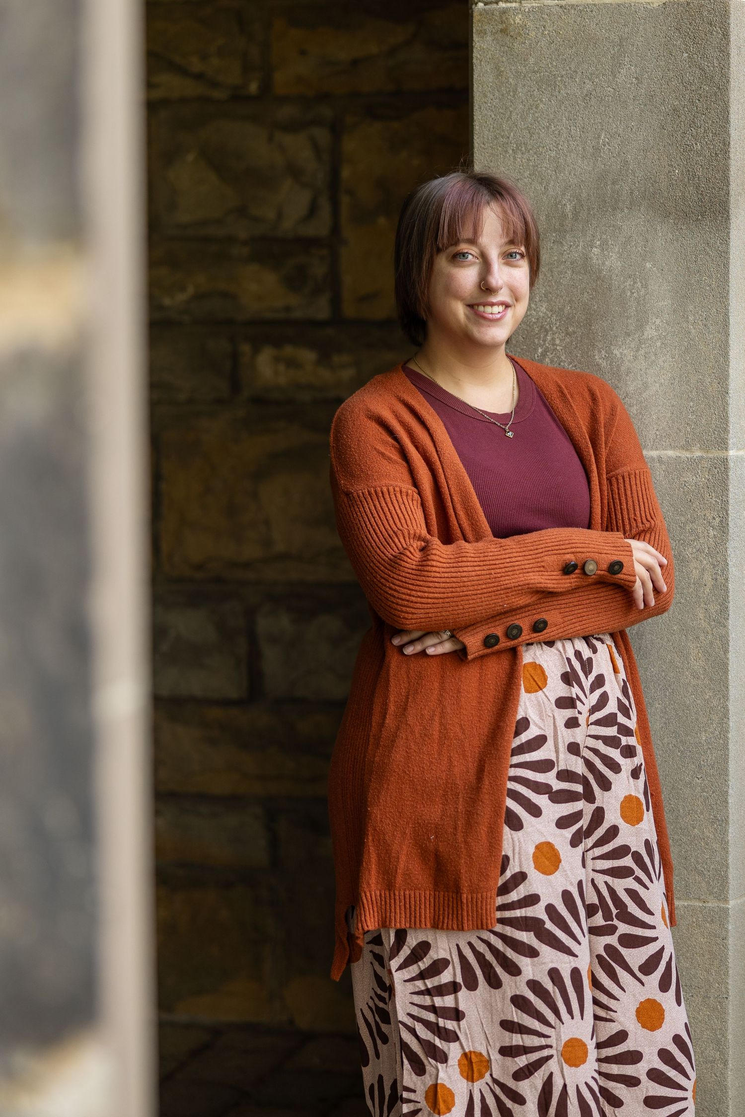 Smiling woman in orange cardigan and floral skirt leaning against stone wall with arms crossed.