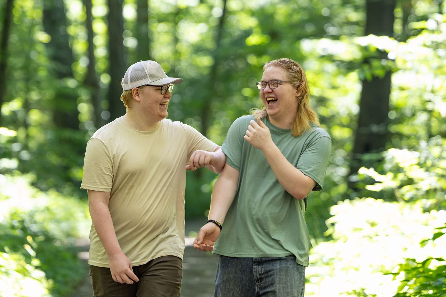 Two friends laughing and walking together on a forest trail surrounded by lush green trees.