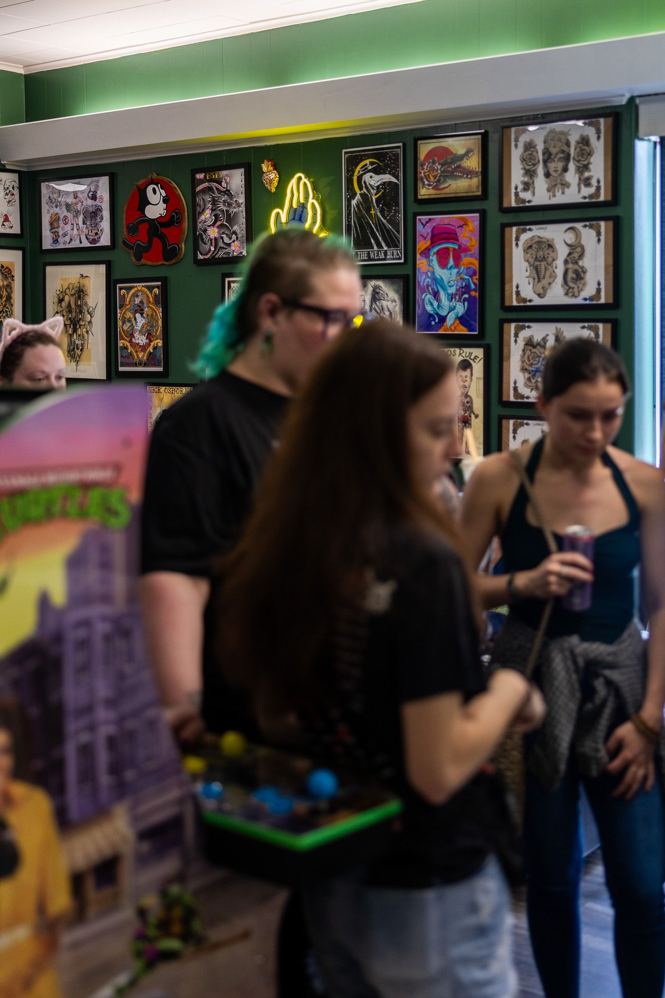 People browsing colorful tattoo flash art displayed on a green wall inside a tattoo shop.