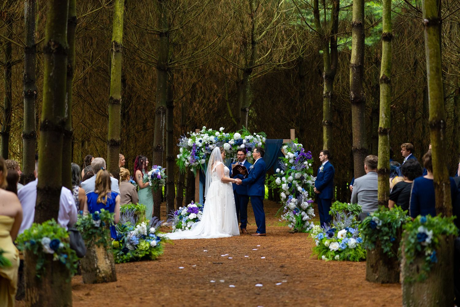 Elegant outdoor forest wedding ceremony with blue floral arch, couple at altar, guests seated along petal-lined aisle.
