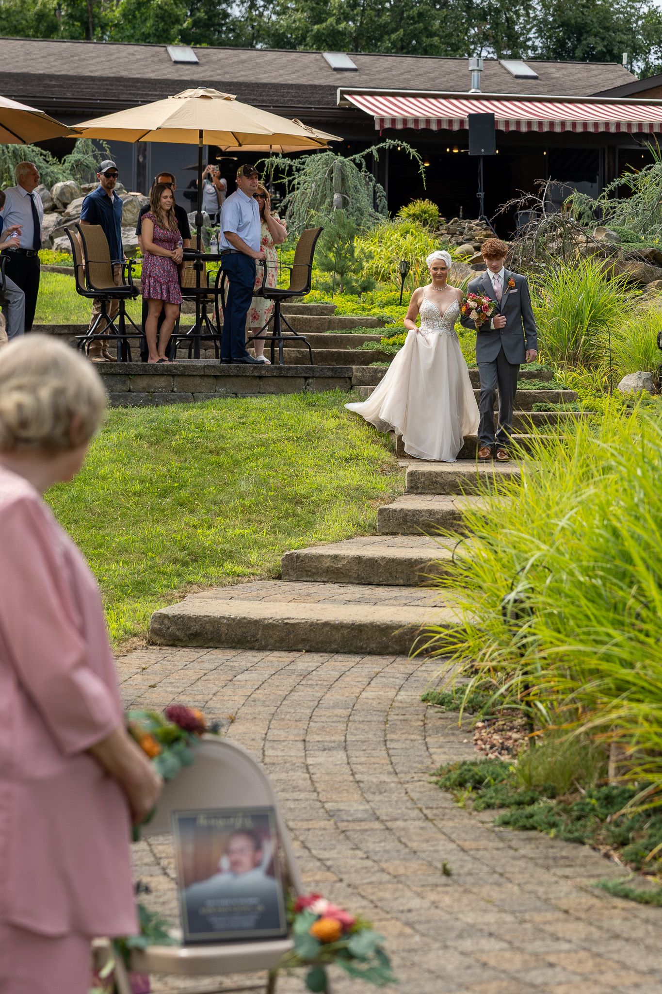 Bride and groom walk down outdoor brick path at garden wedding venue surrounded by lush greenery and guests.