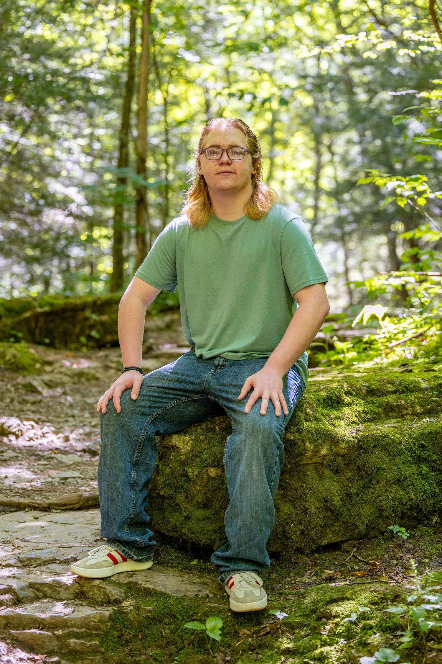 Young man with long blonde hair and glasses sits on mossy rocks in a lush green forest wearing a teal t-shirt.