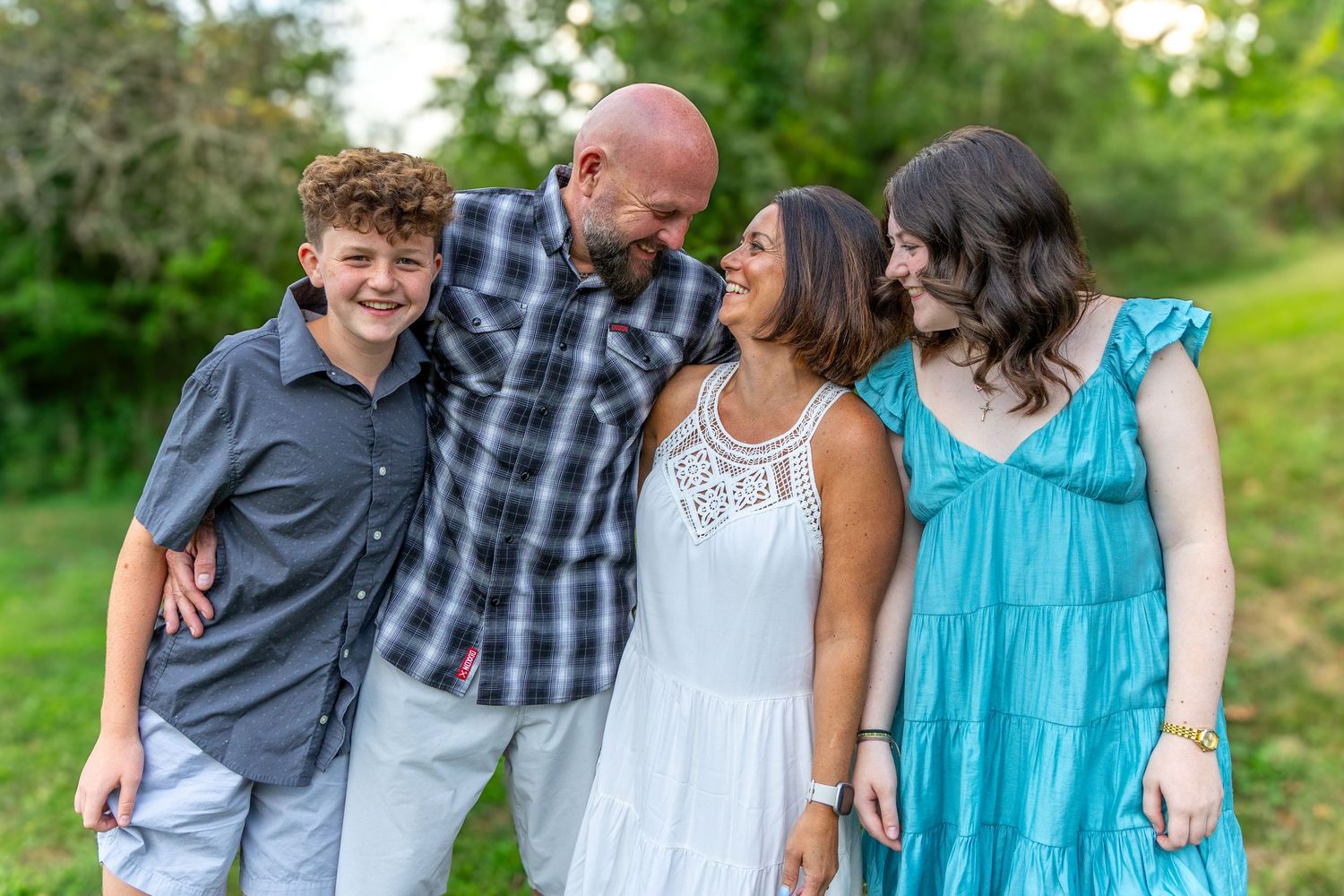 A joyful family of four laughing together outdoors, with lush green trees in the background on a sunny day.