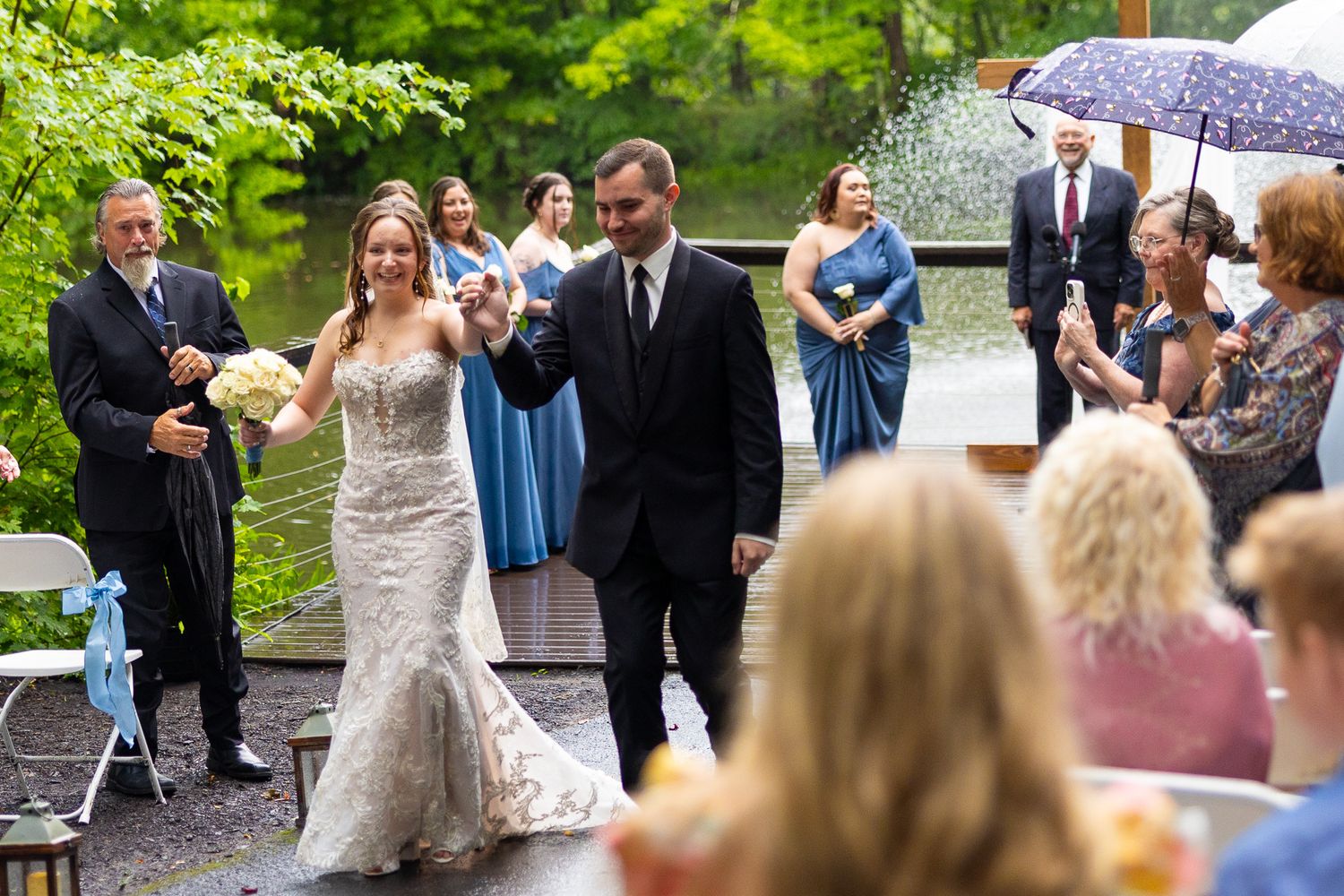 Newlyweds walk joyfully down aisle after rainy outdoor wedding ceremony as guests celebrate.