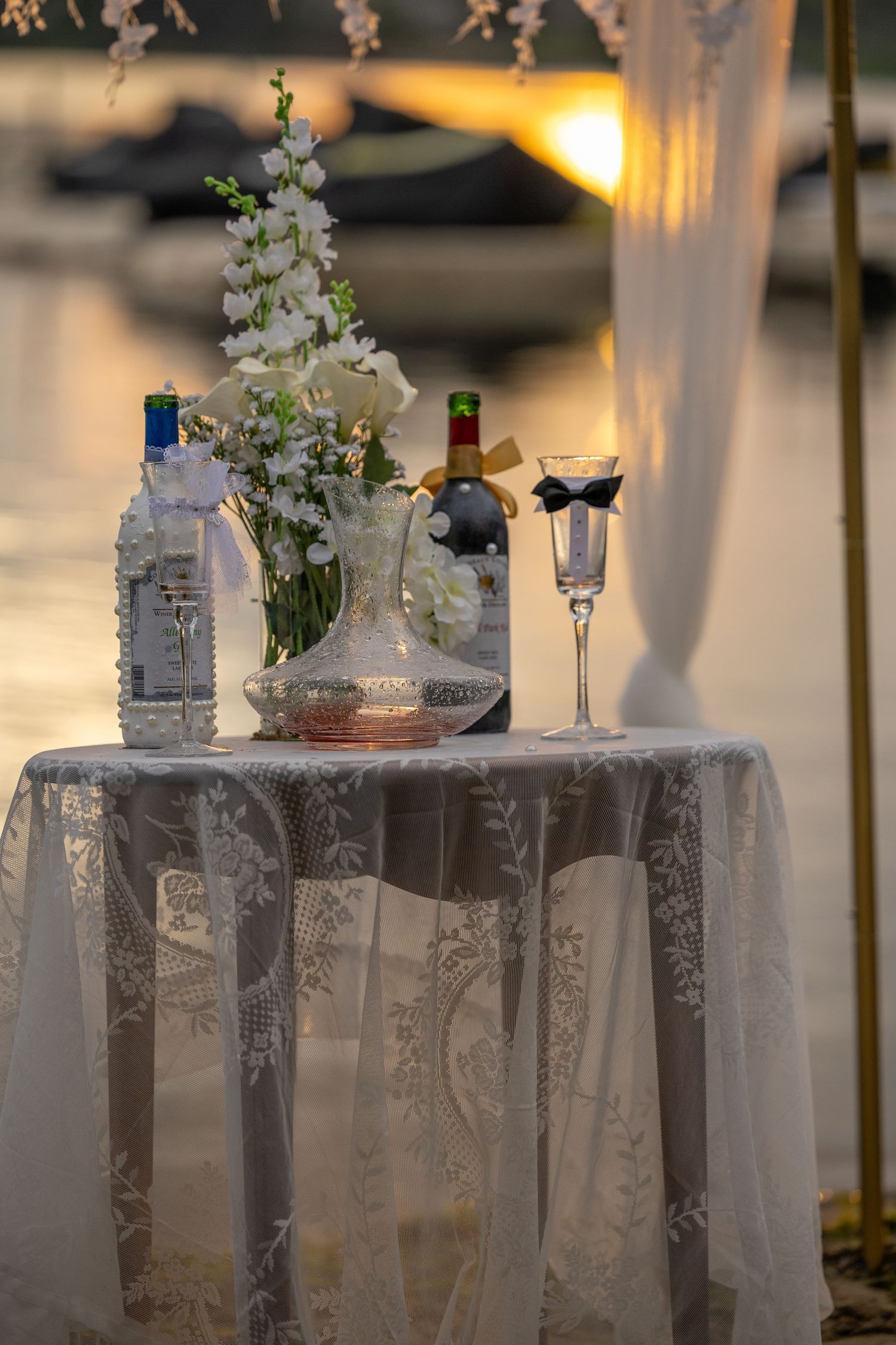 Elegant wedding table with lace cloth, wine bottles, floral arrangement, and decorated champagne glass at sunset by the water.