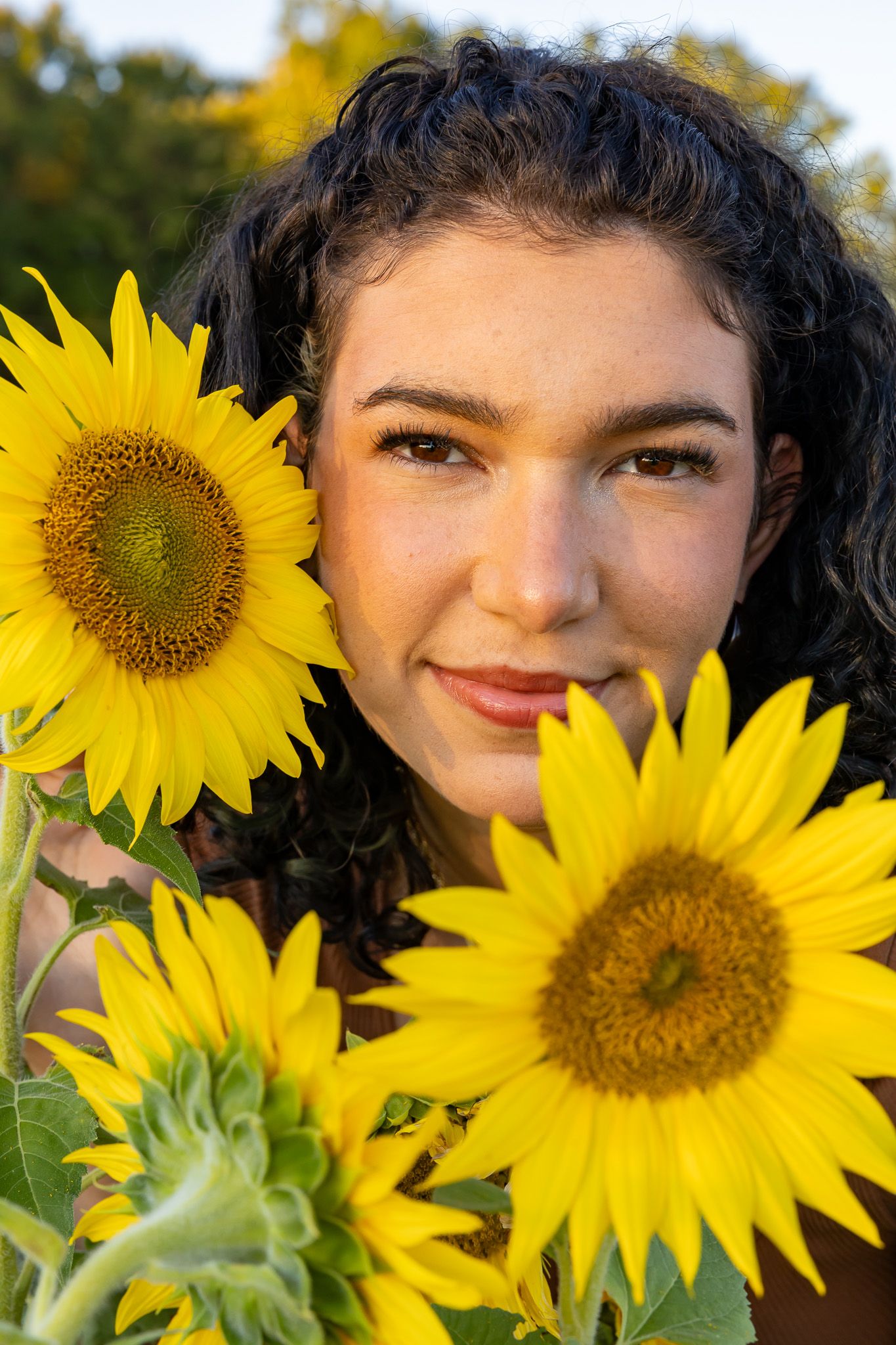 Young woman with curly dark hair smiling among bright yellow sunflowers in a sunny field.