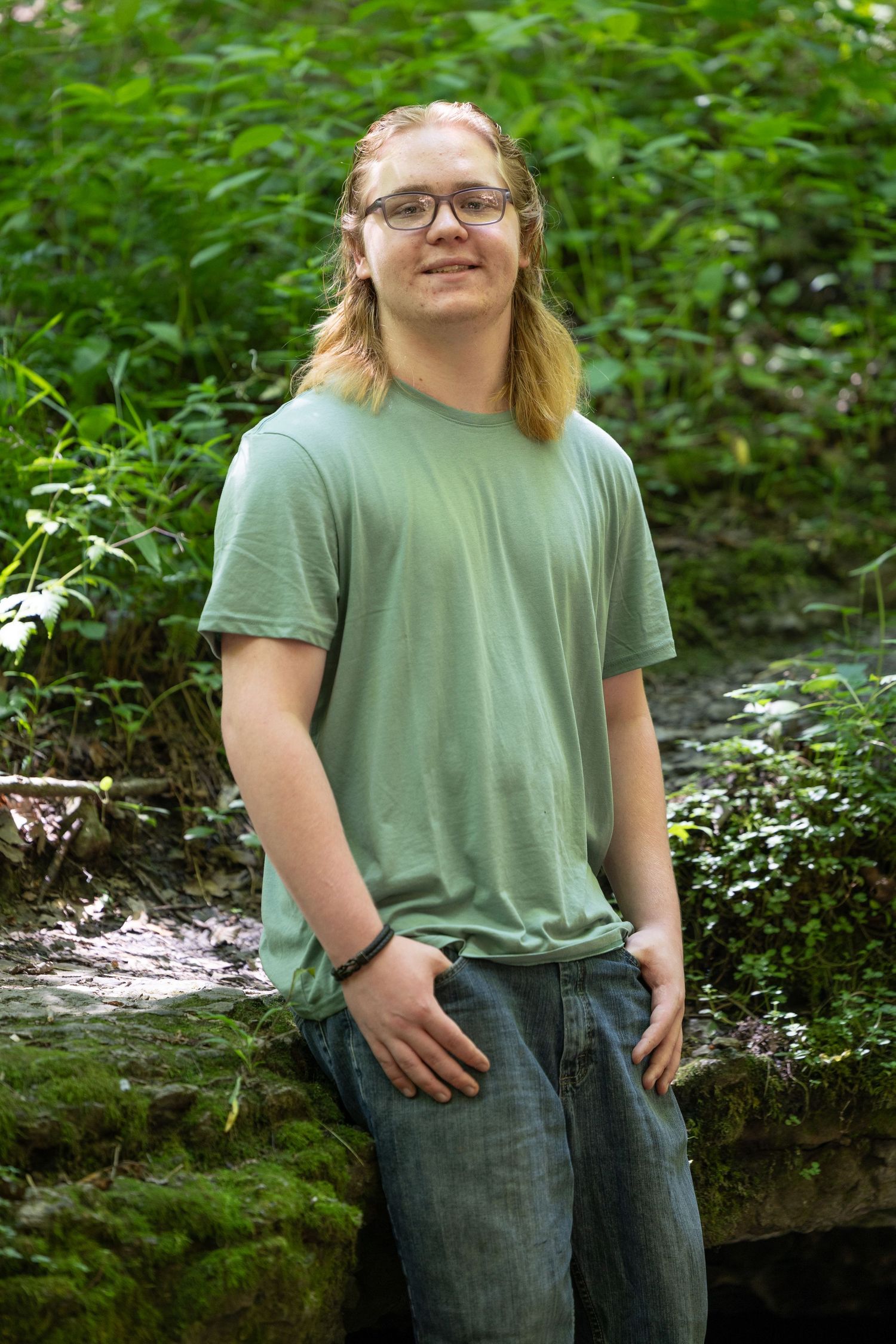 Young man with long blonde hair and glasses wearing a green t-shirt, leaning against a mossy wall in a forest.