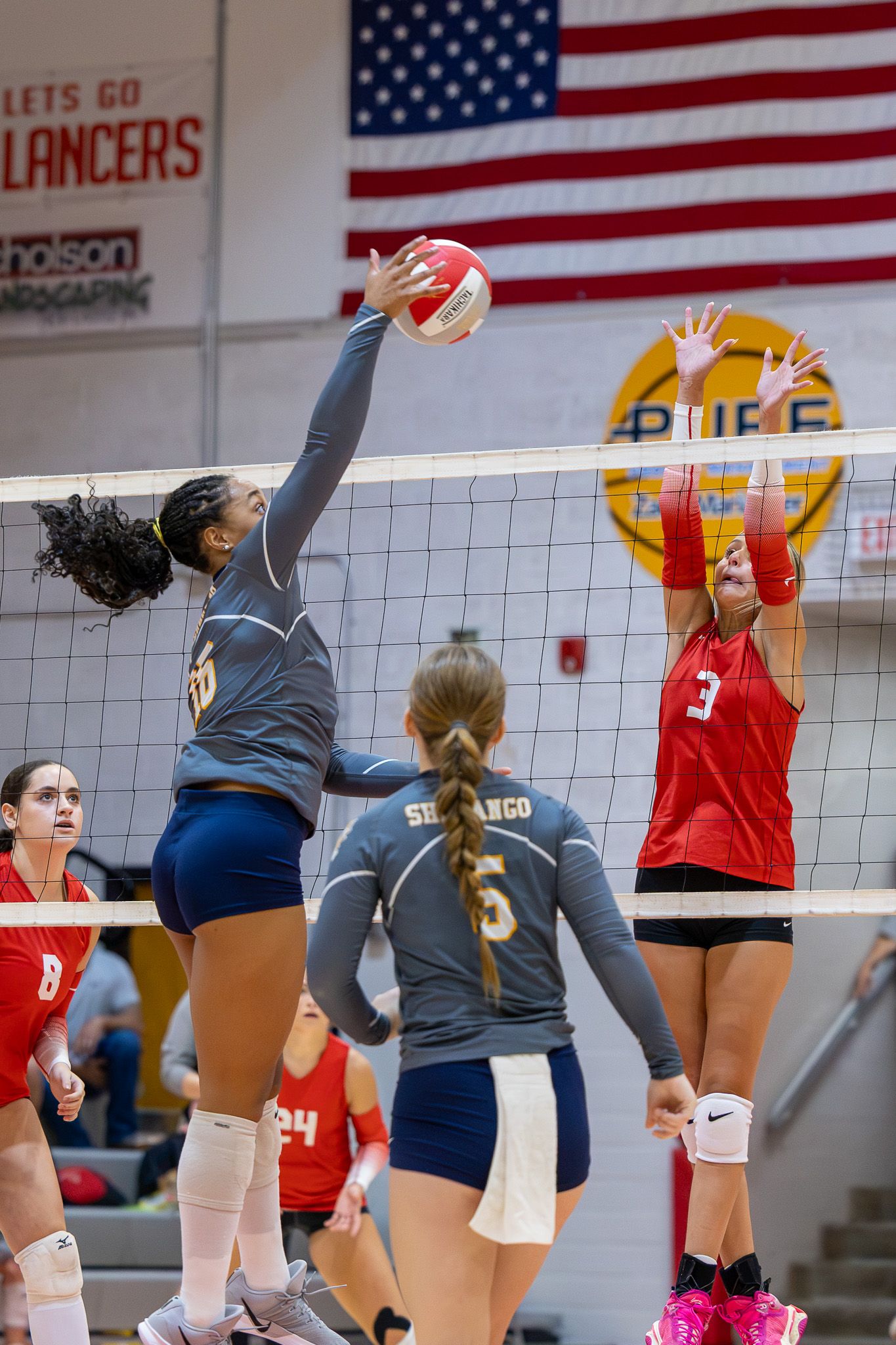 Female volleyball players compete at the net during an indoor high school match, with one player spiking the ball.