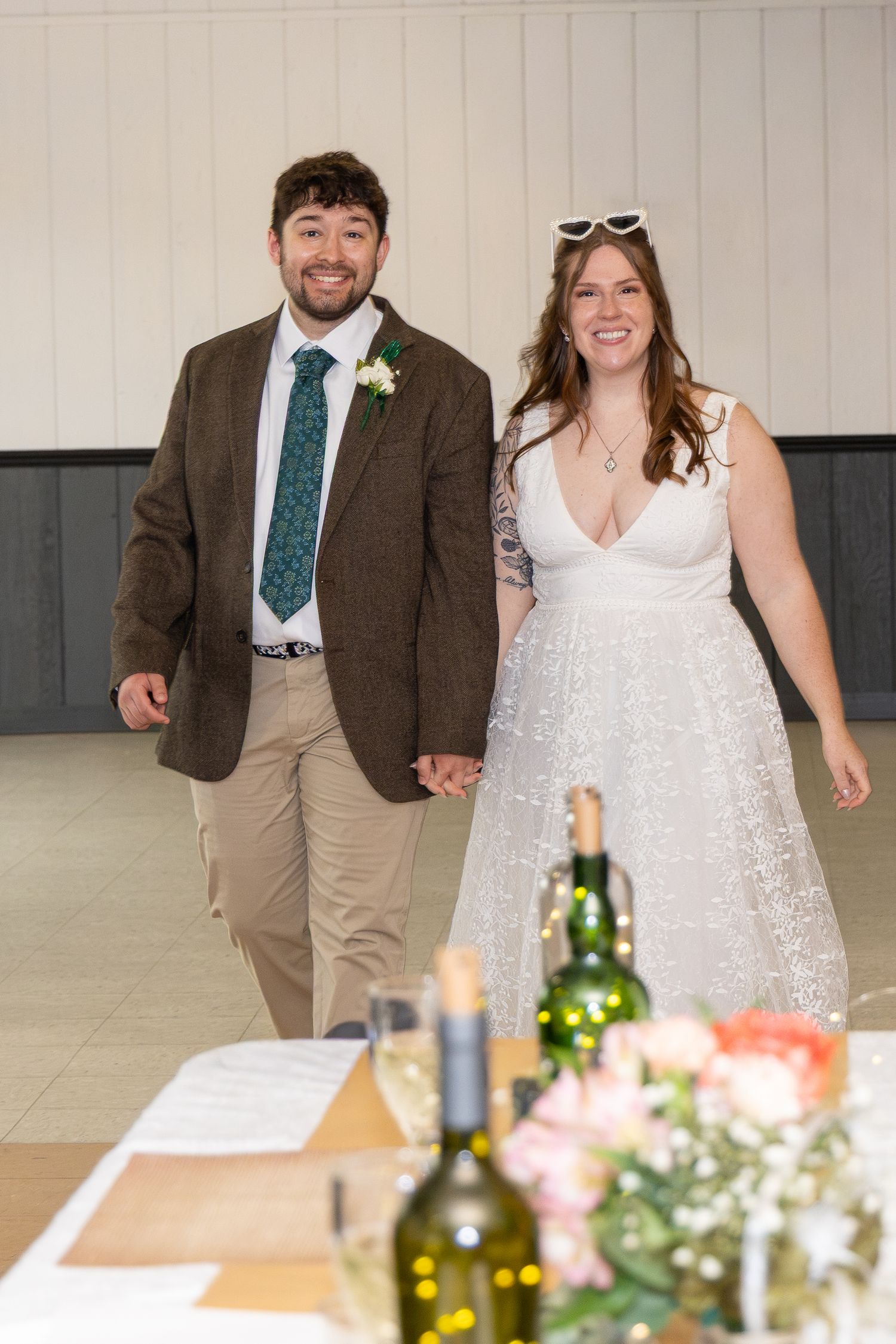Happy newlyweds holding hands at their wedding reception, groom in brown suit with green tie, bride in white sparkly dress.