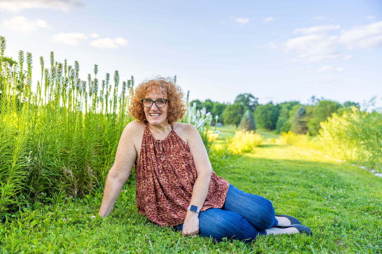Woman with curly red hair sitting on green grass beside tall plants in a sunny outdoor field.