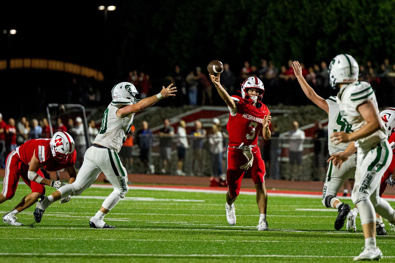 High school football quarterback throws a pass under pressure during a night game under bright stadium lights.