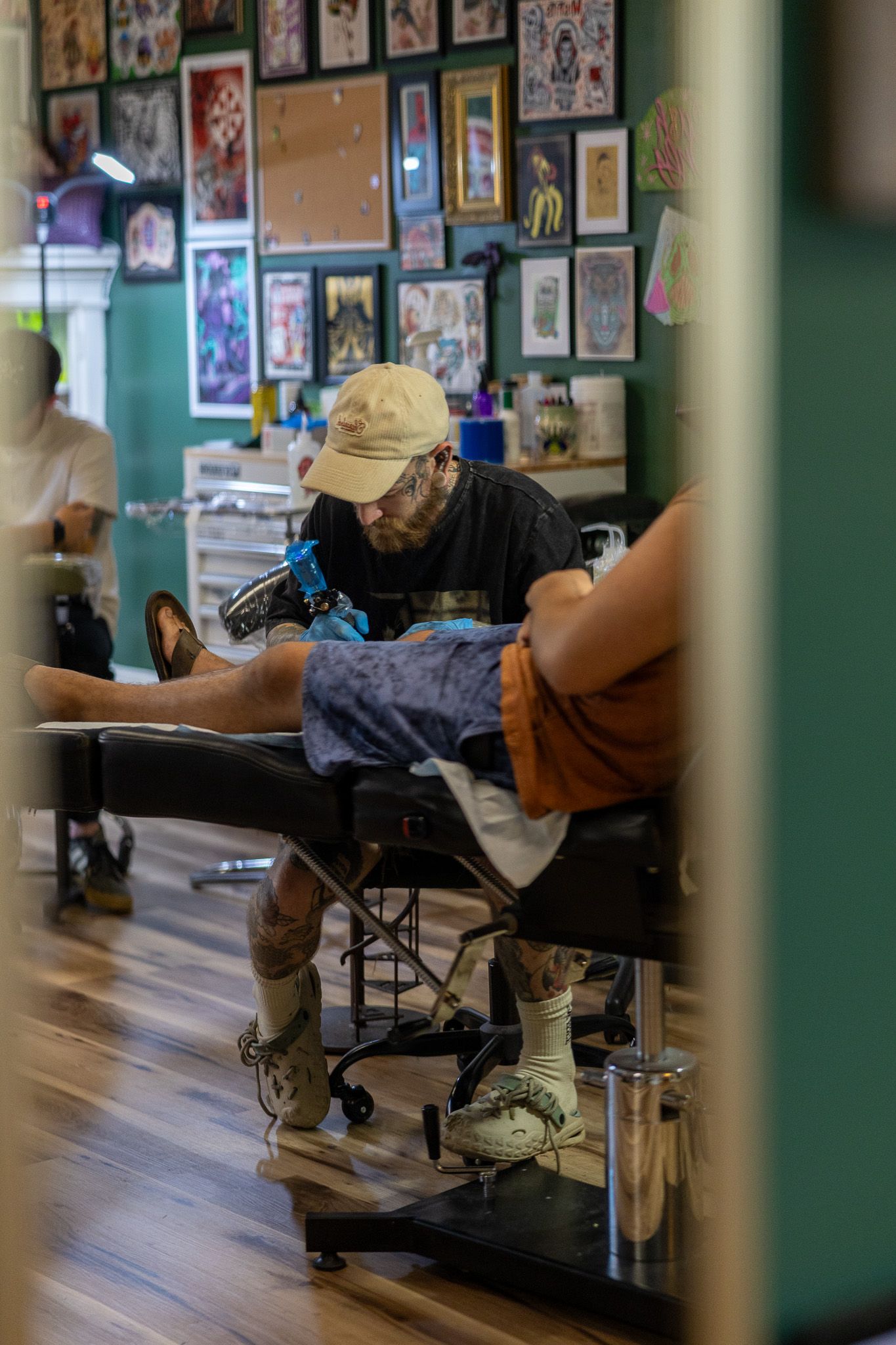 Tattoo artist in cap working on client's leg in a colorful studio with flash art covering teal walls.