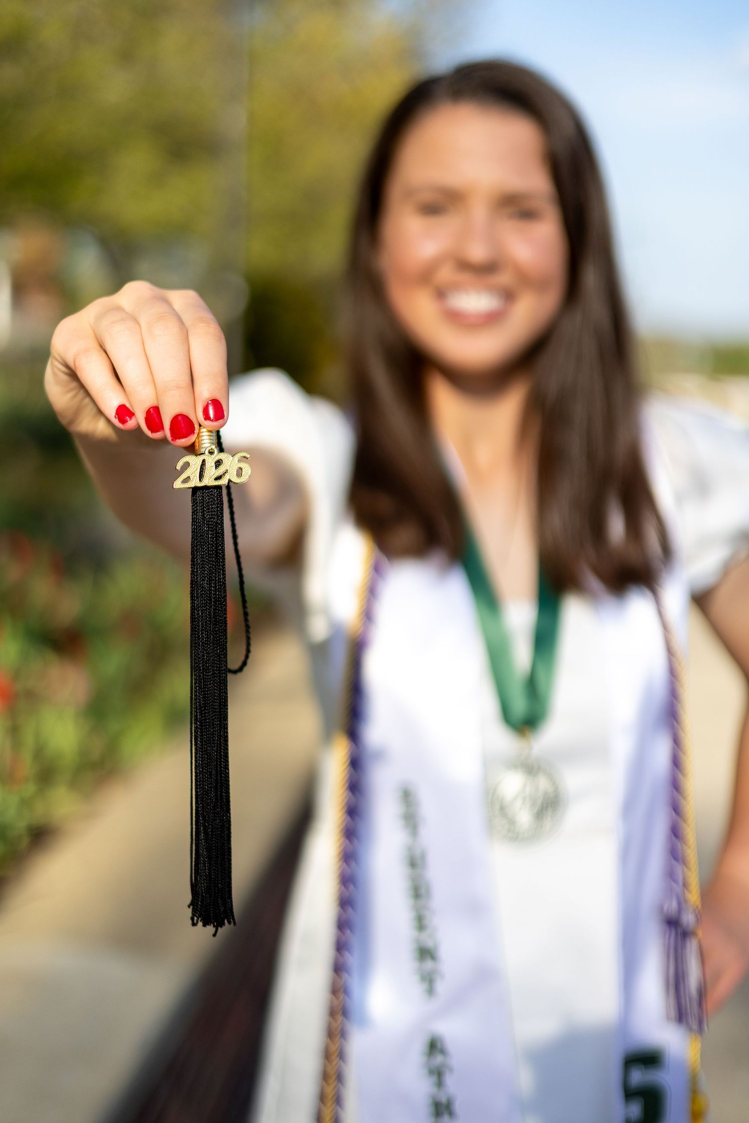 Smiling graduate in white dress holds 2026 tassel charm toward camera outdoors.