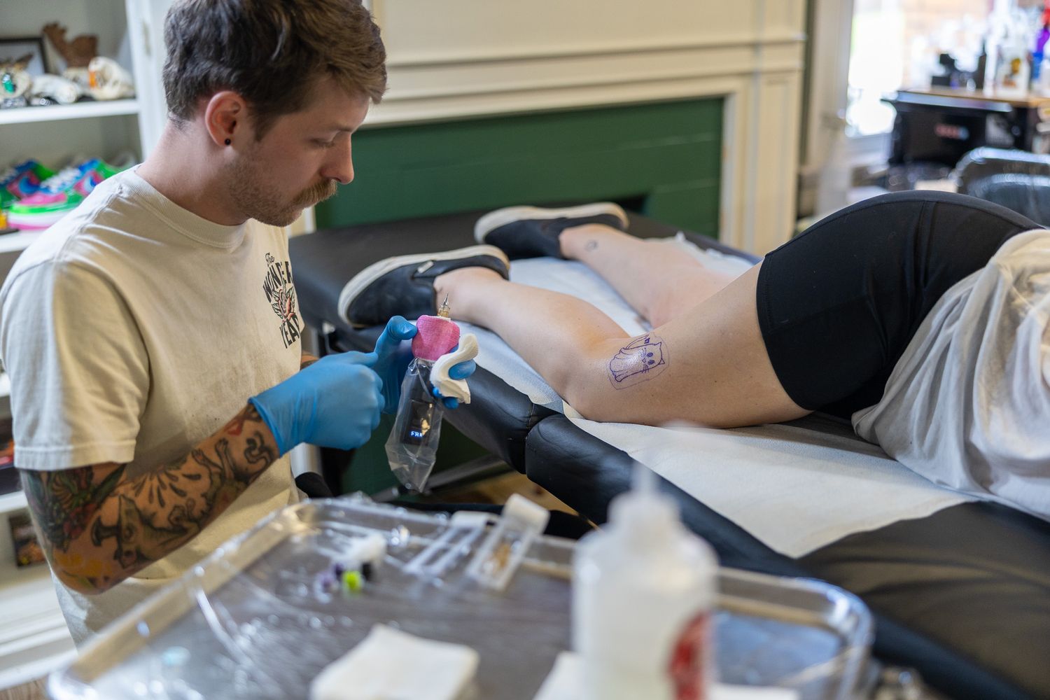Tattoo artist with sleeve tattoos working on a clients leg in a bright tattoo parlor.