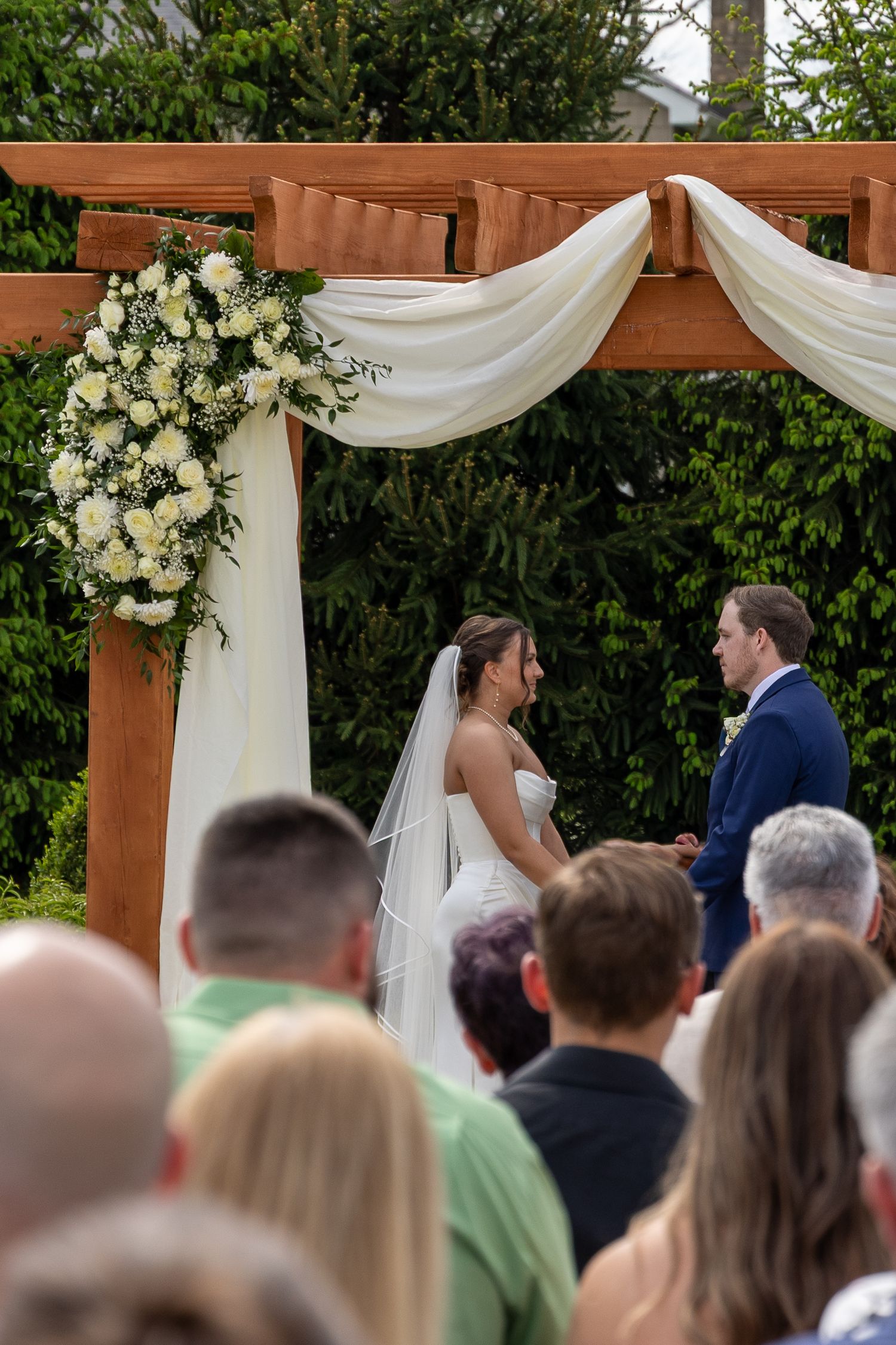 Bride and groom exchange vows under a wooden arch with white draping and floral arrangements at an outdoor wedding ceremony.