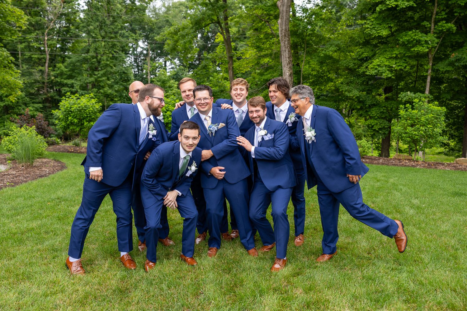 Groomsmen in navy blue suits laugh and playfully pose together in a lush green outdoor setting.