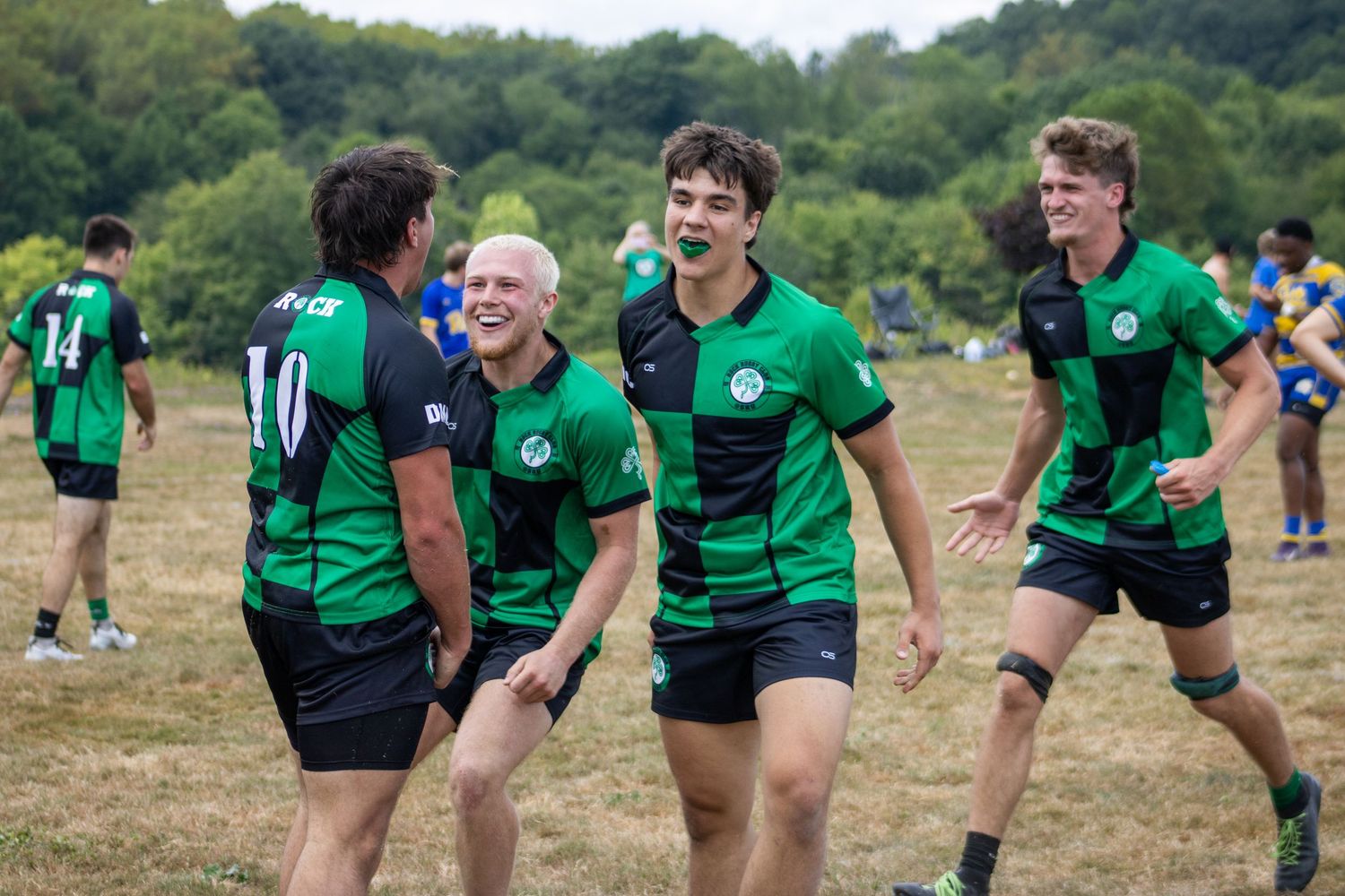 Four smiling rugby players in green uniforms celebrate together after a successful play on the field.
