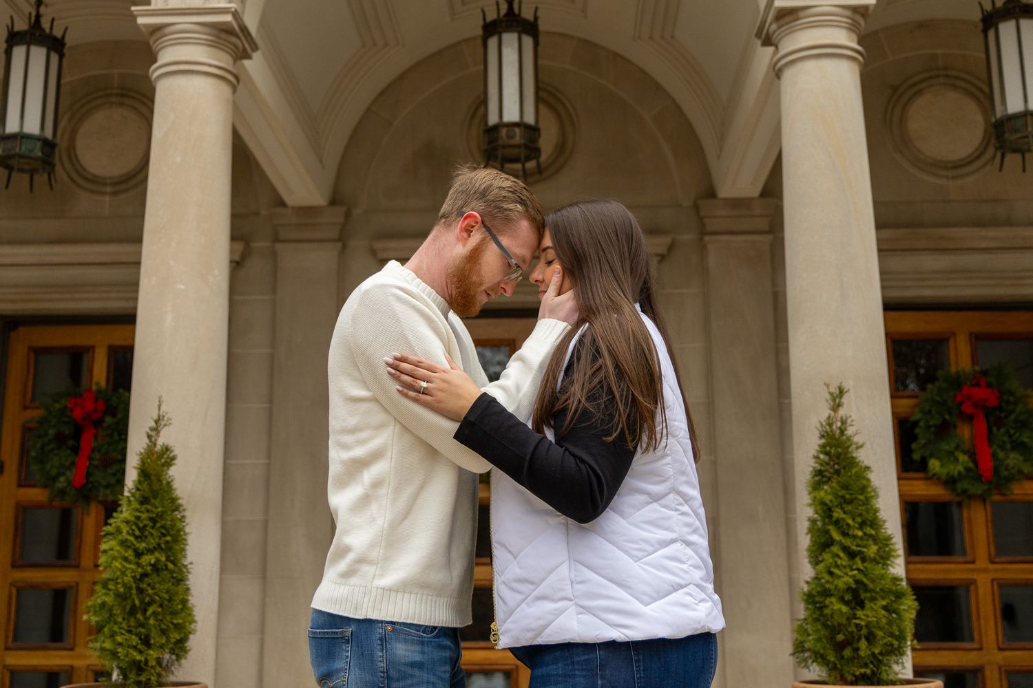 A couple sharing a romantic kiss in front of an elegant building decorated with Christmas wreaths.