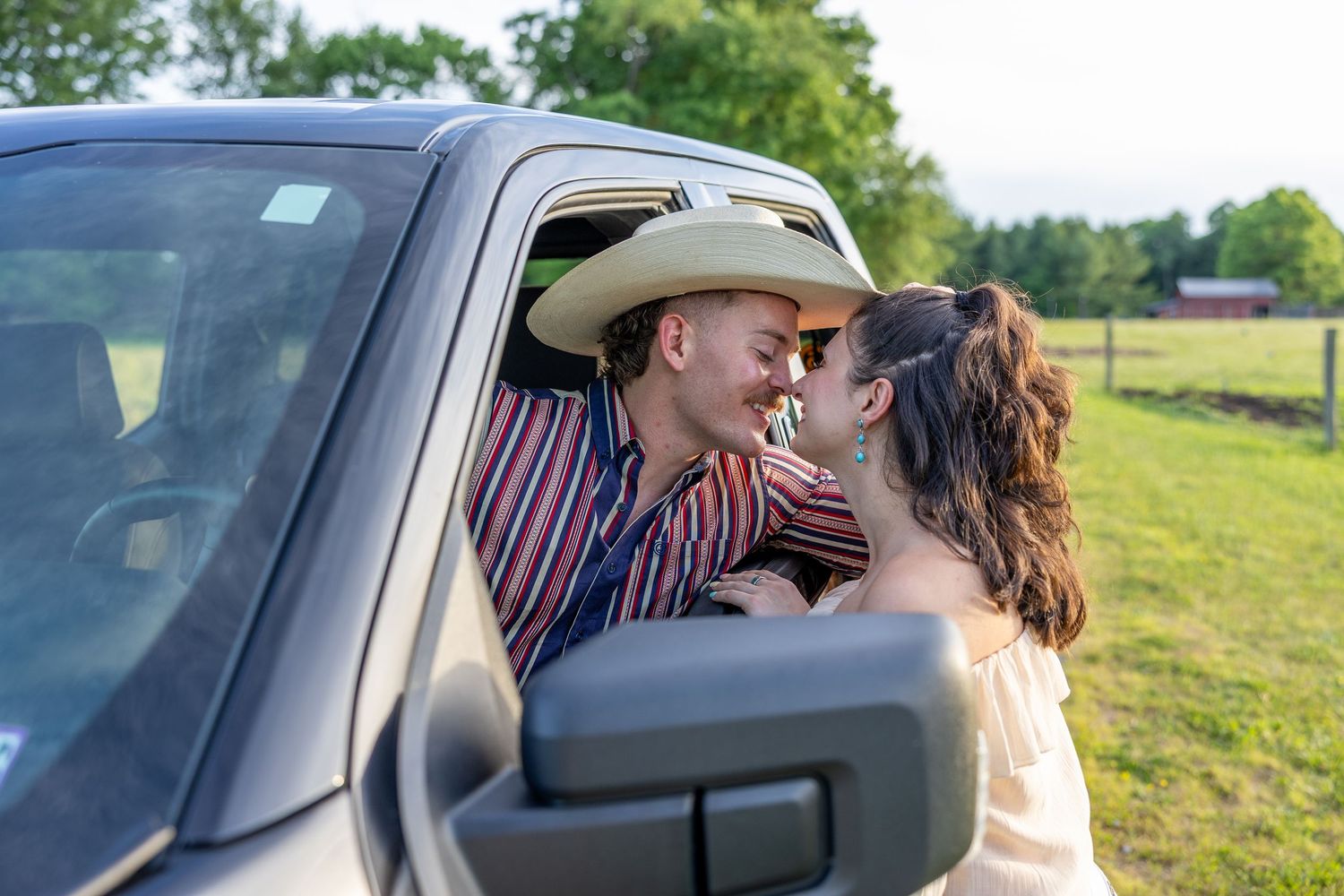 A couple sharing a romantic moment near a truck window in a sunny green field, man wearing a cowboy hat.