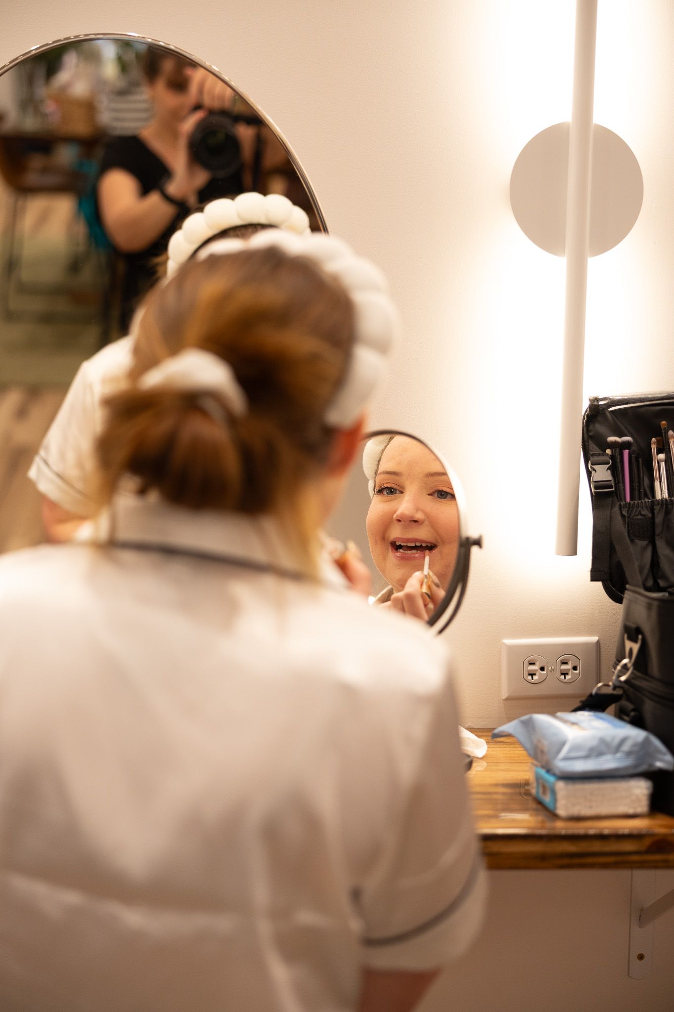 Young woman in white uniform applies lipstick while smiling at a small mirror at a vanity dressing table.