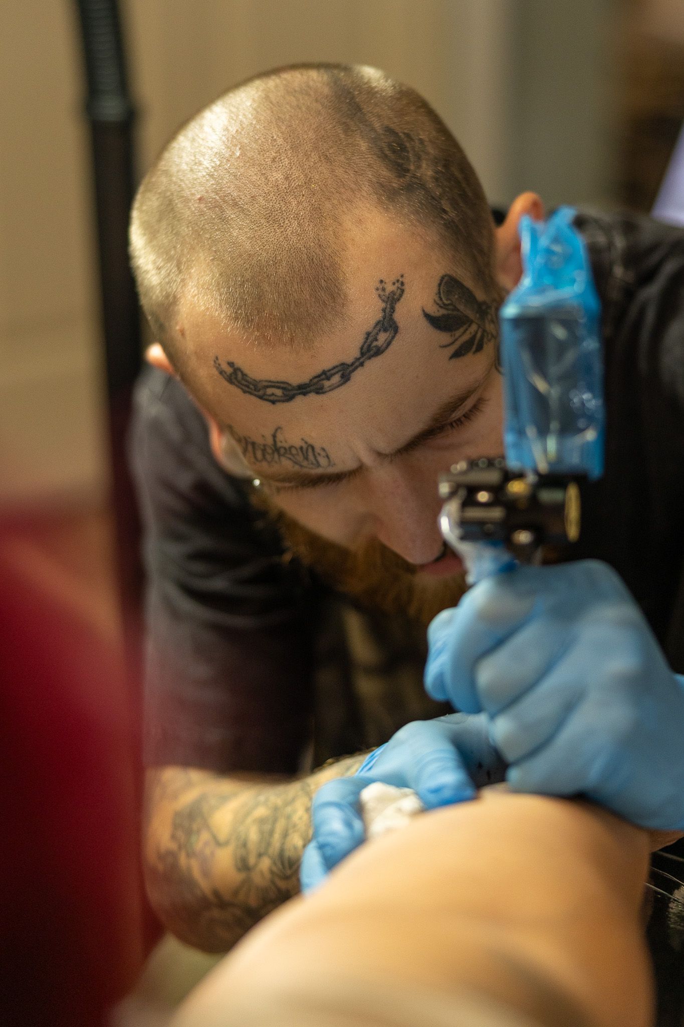Heavily tattooed artist with face tattoos carefully applies tattoo using machine while wearing blue gloves.