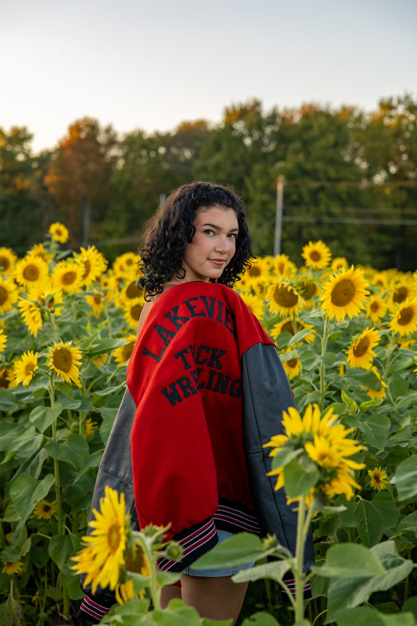 Young woman in red Lakeview wrestling varsity jacket poses in a vibrant sunflower field at golden hour.
