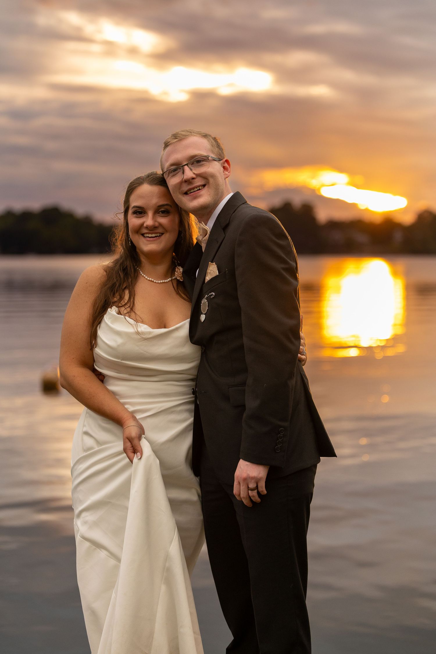 Bride and groom smiling together by a lake at sunset, bride in white gown and groom in dark suit.