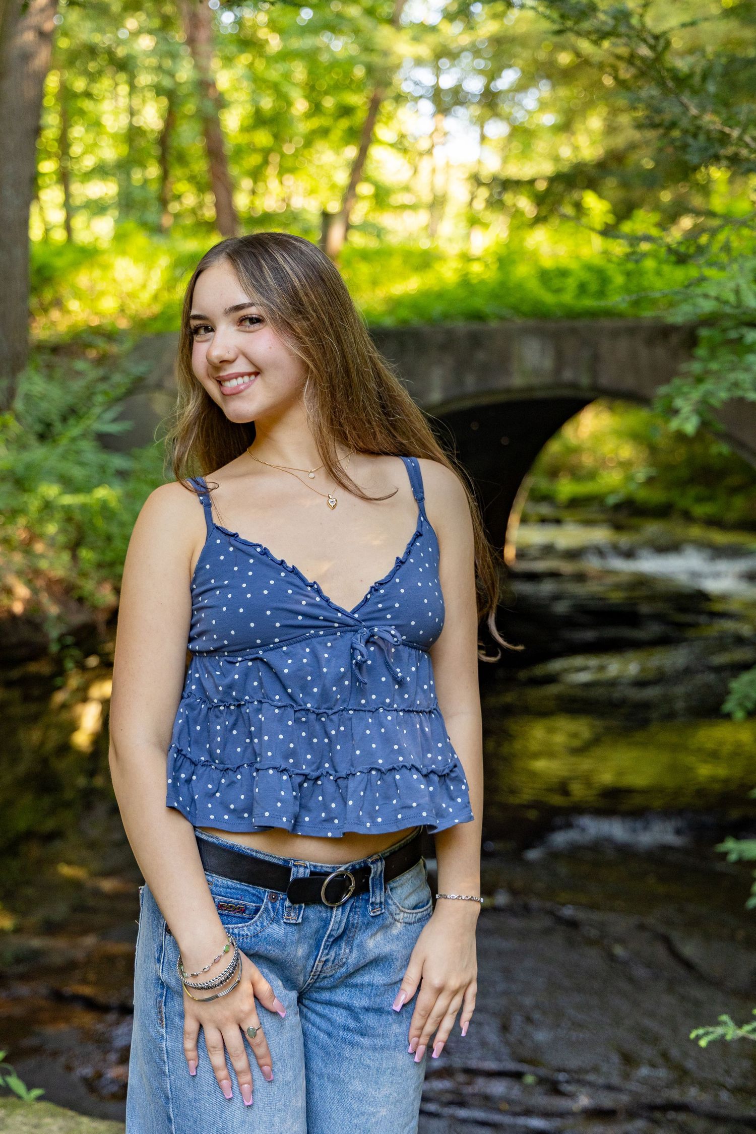 Young woman in blue polka dot top and jeans smiling outdoors near a stone bridge and stream in a lush forest.