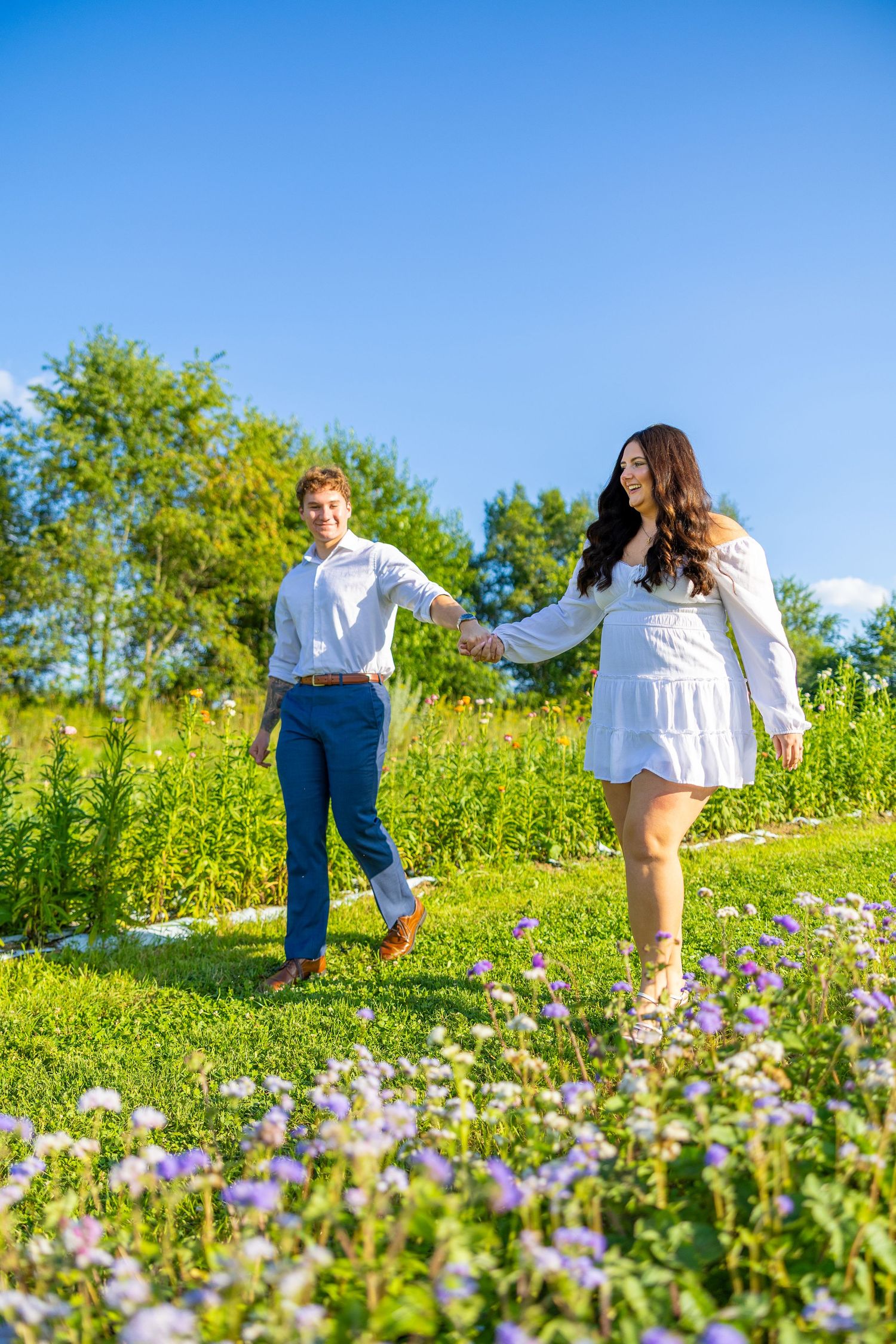 Happy couple holds hands walking through wildflower meadow on sunny day during engagement photo session.